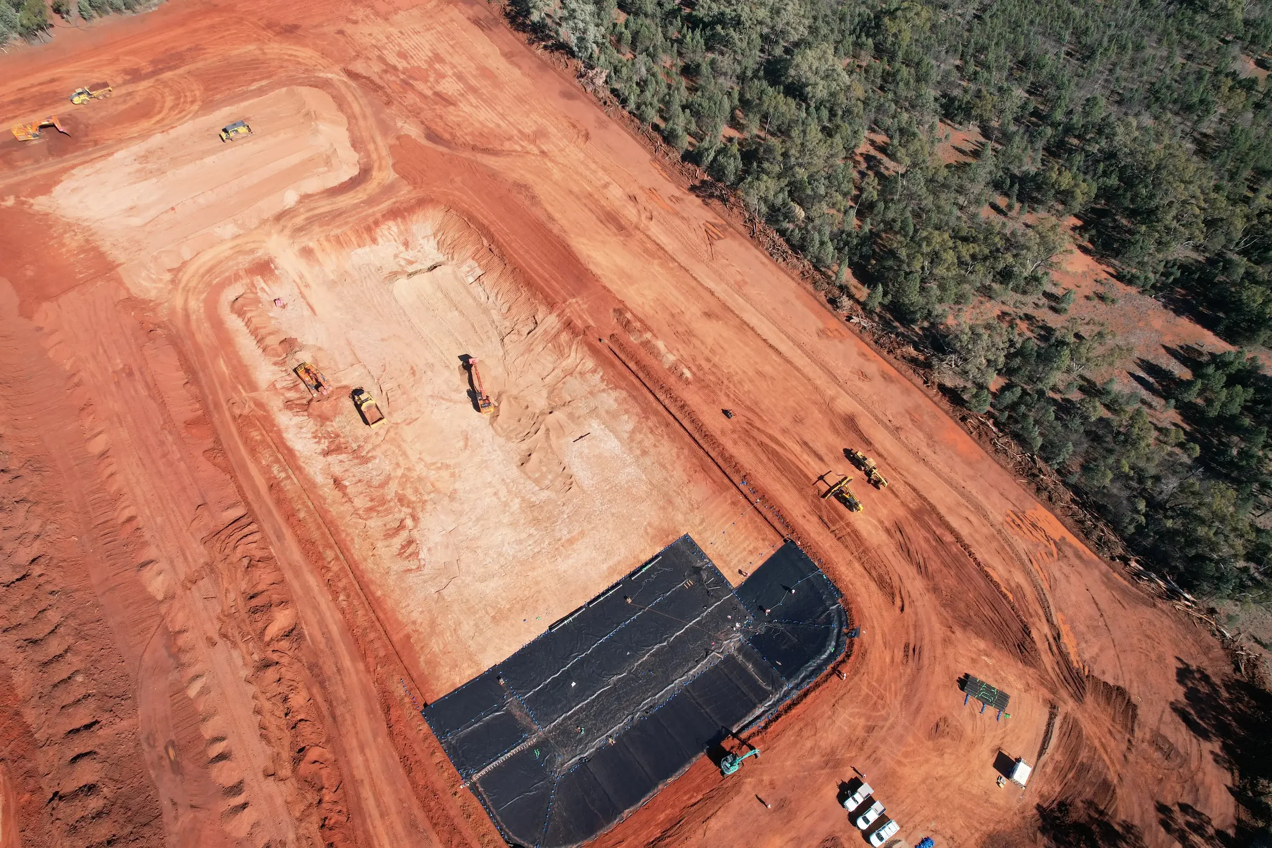 Aerial view of a construction site with excavators, trucks, and a partially built structure on red soil, surrounded by a forest.