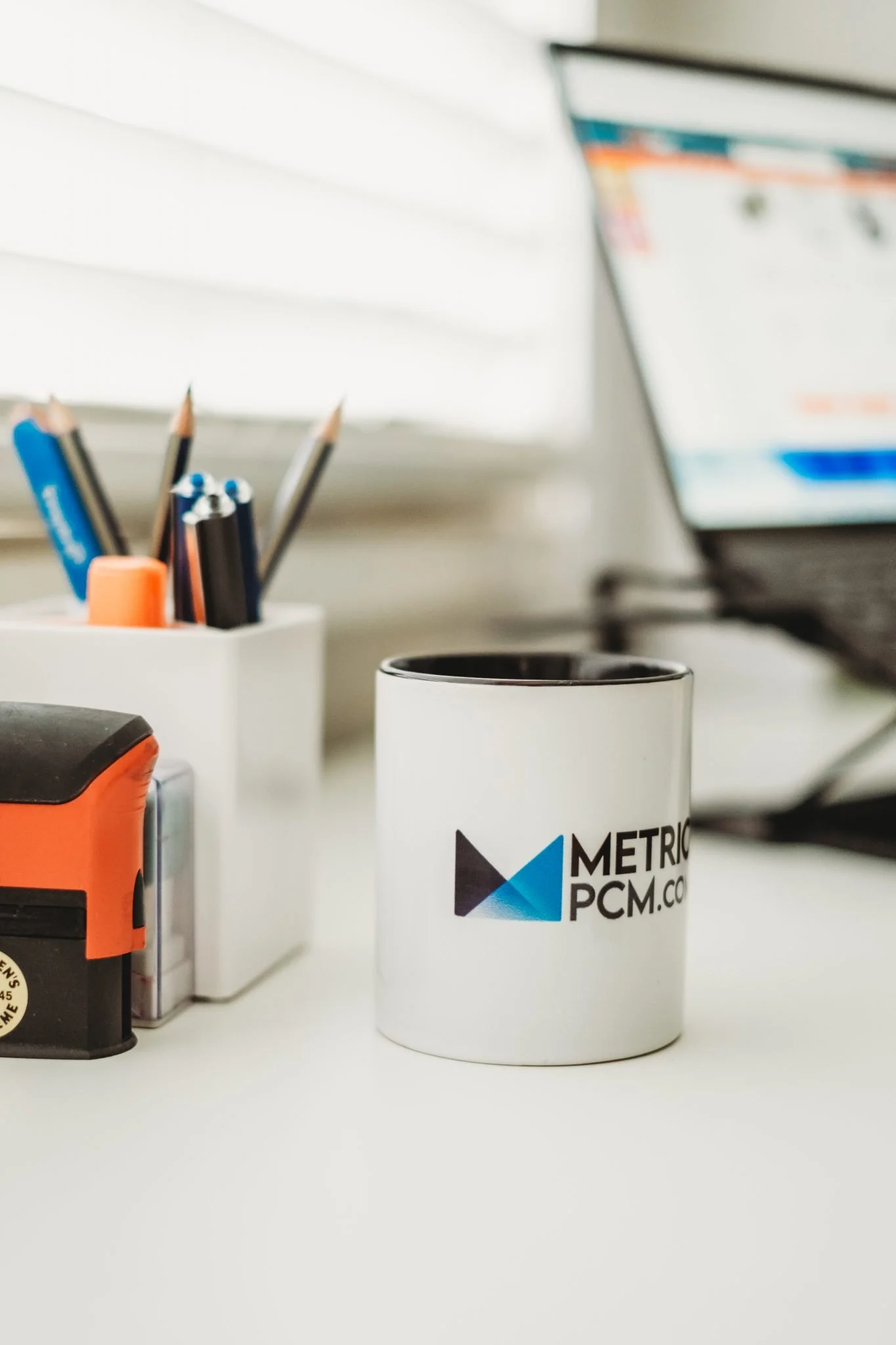 White coffee mug with 'METRIC PCM.co' logo on a white desk, surrounded by office supplies and a computer in the background.