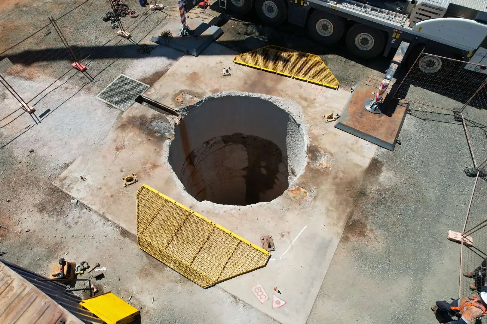Construction site with a large hole in the concrete, surrounded by safety barriers and construction equipment.