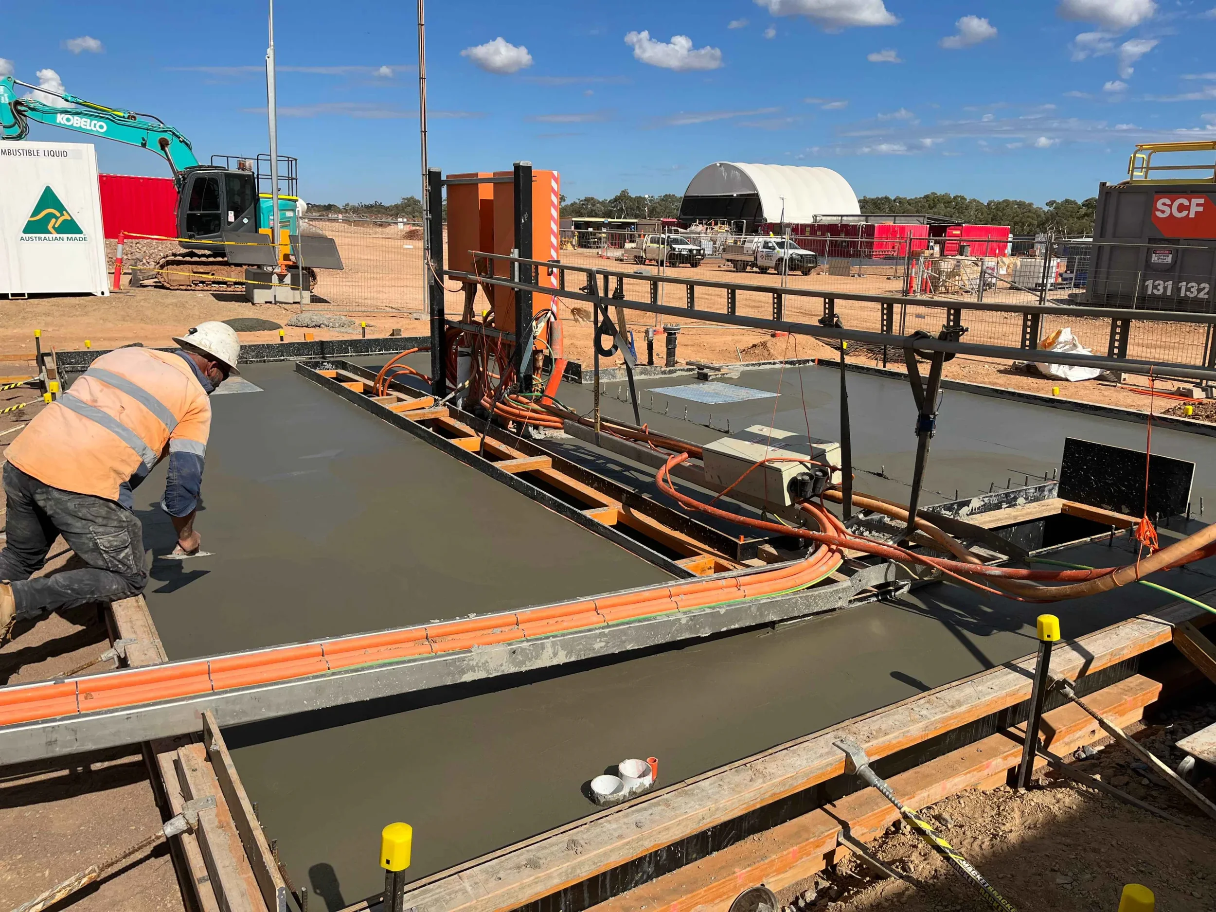 Construction workers pouring concrete into a foundation at a building site, with construction equipment, vehicles, and a blue sky in the background.