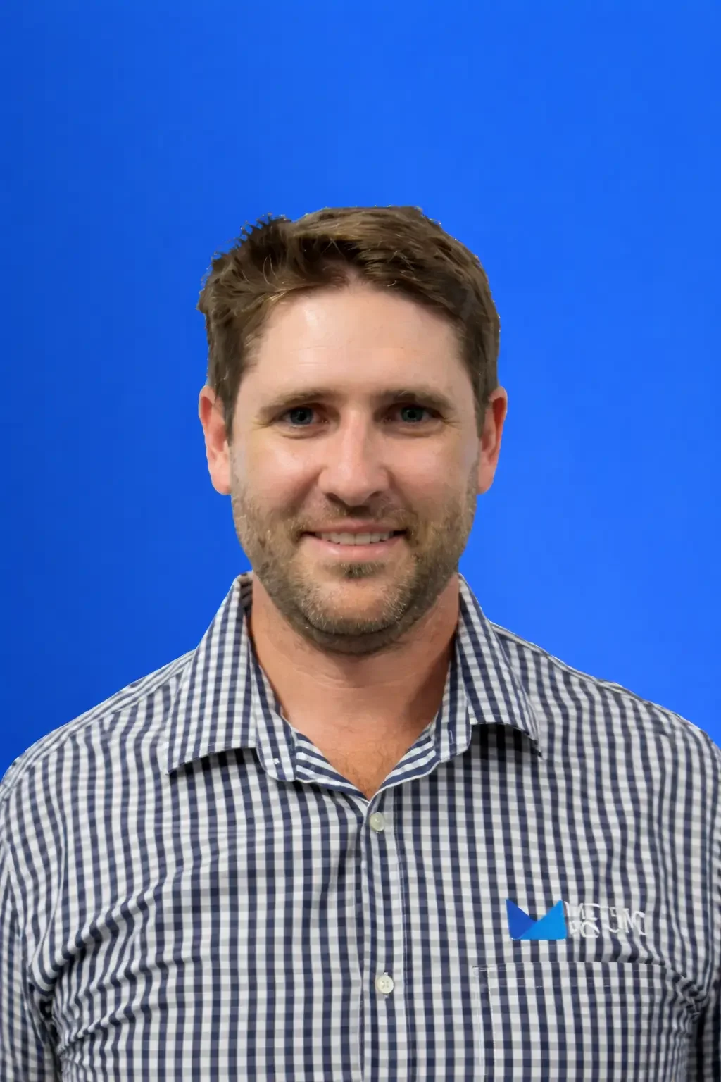 Portrait of a smiling man with short brown hair and a beard, wearing a checkered shirt with a company logo, against a solid blue background.