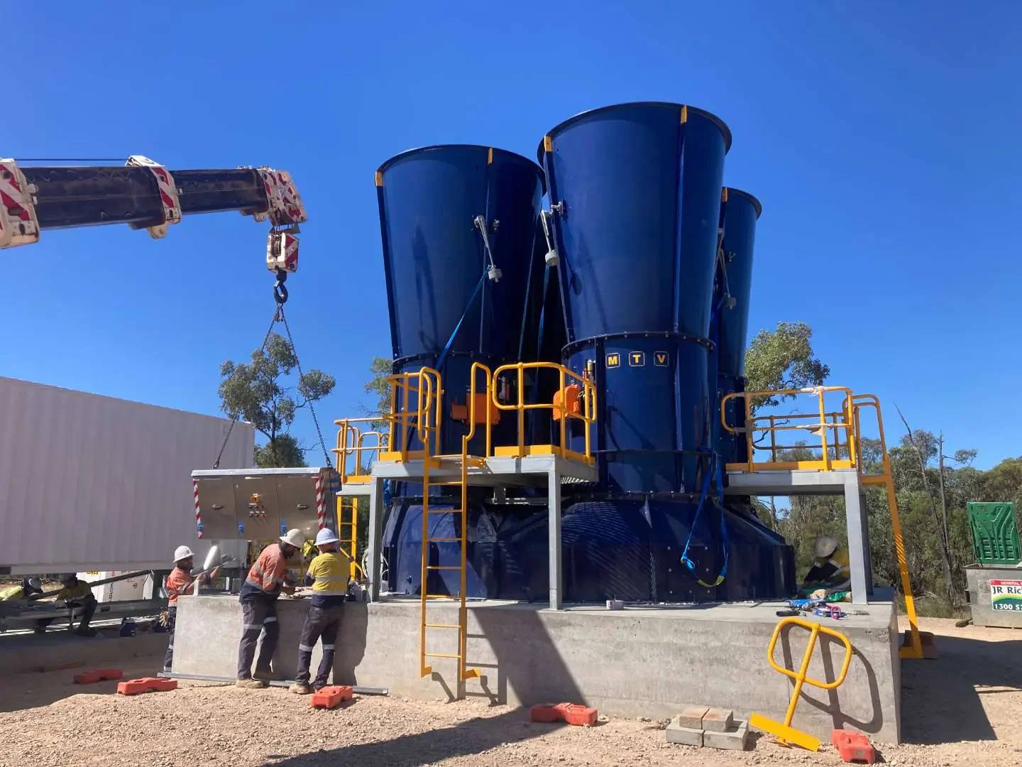 Workers in safety gear installing large blue industrial equipment on a concrete platform while a crane assists with lifting parts, outdoors under a clear blue sky.