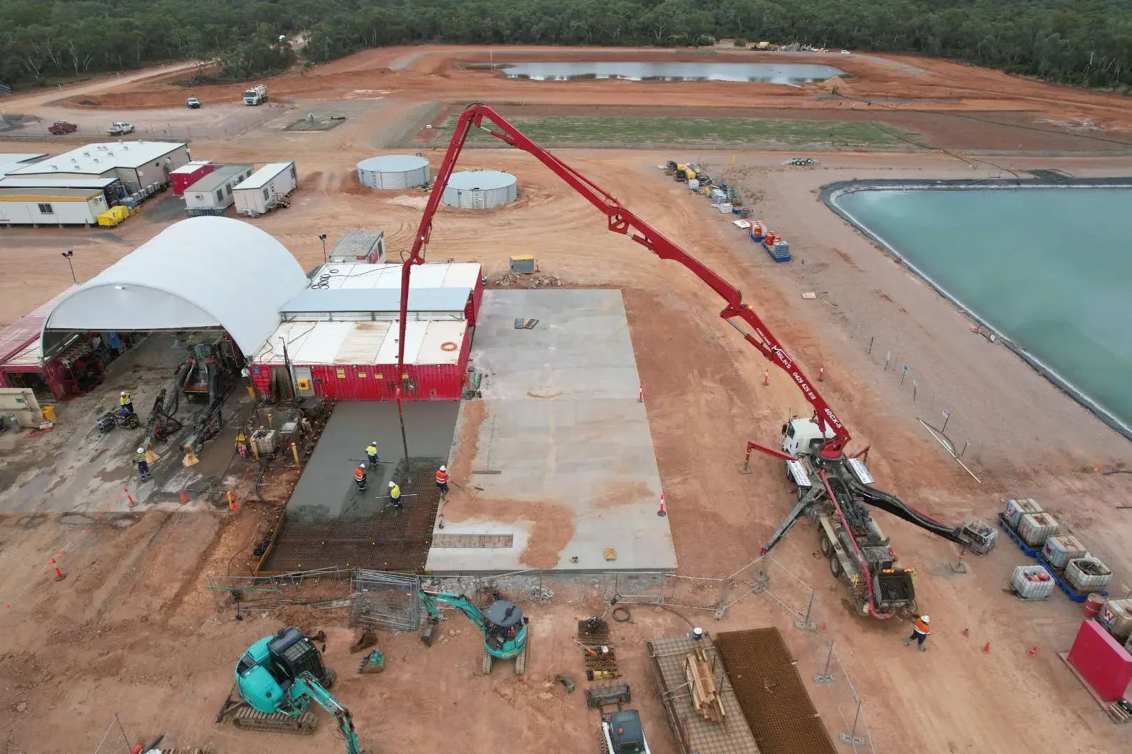 Construction site with workers, pouring concrete, and a concrete pump truck, near water tanks and ponds.