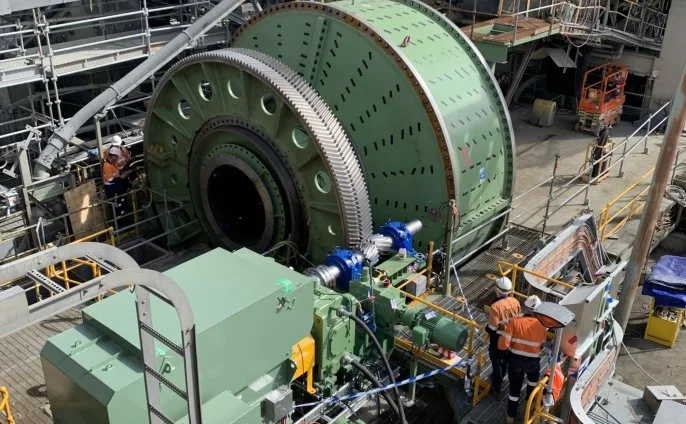 Workers in safety vests and helmets working on large machinery at an industrial site, with a massive green rotating component and various equipment surrounding it.