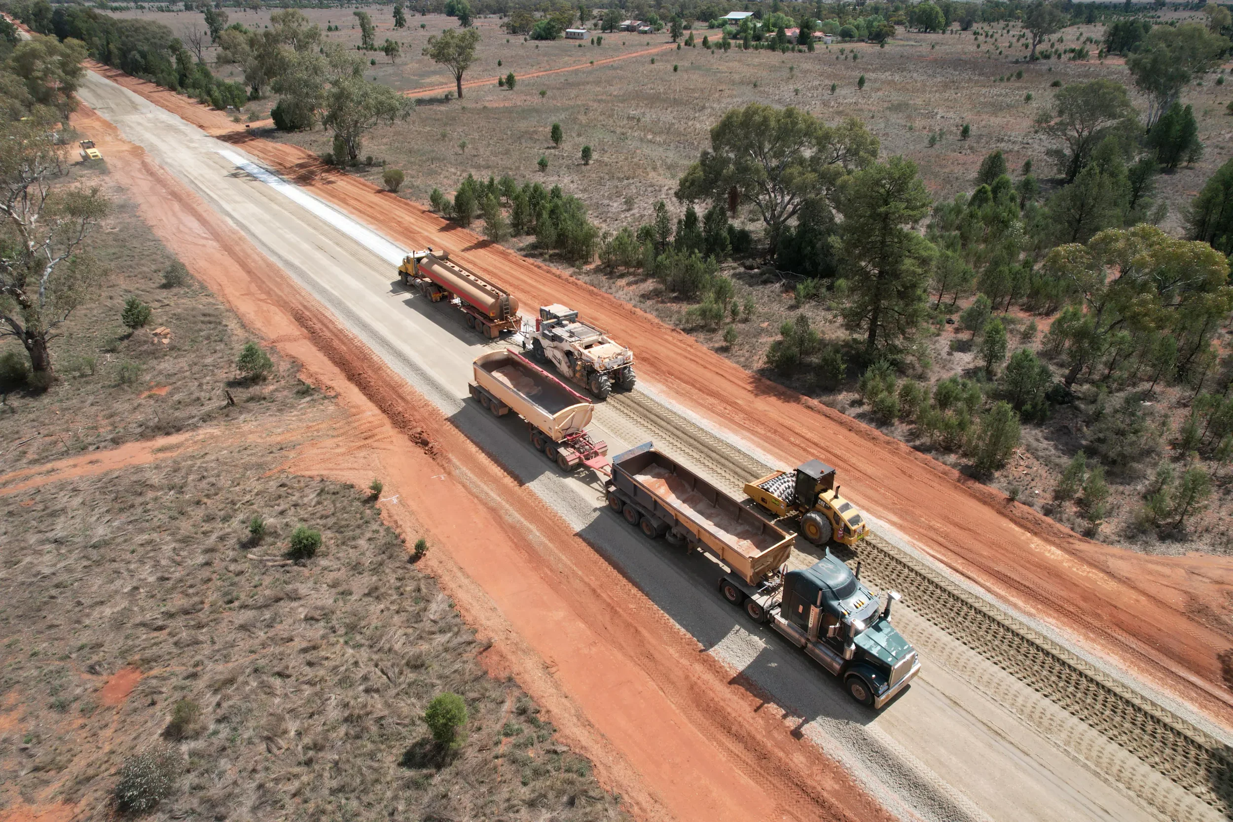 Aerial view of a road construction site with multiple construction vehicles laying asphalt on a rural road surrounded by trees and open land.