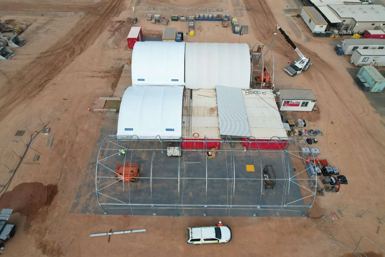 Aerial view of a construction site with temporary structures, including a greenhouse frame, tents, and mobile offices, on reddish dirt ground, with construction vehicles, materials, and equipment parked around.