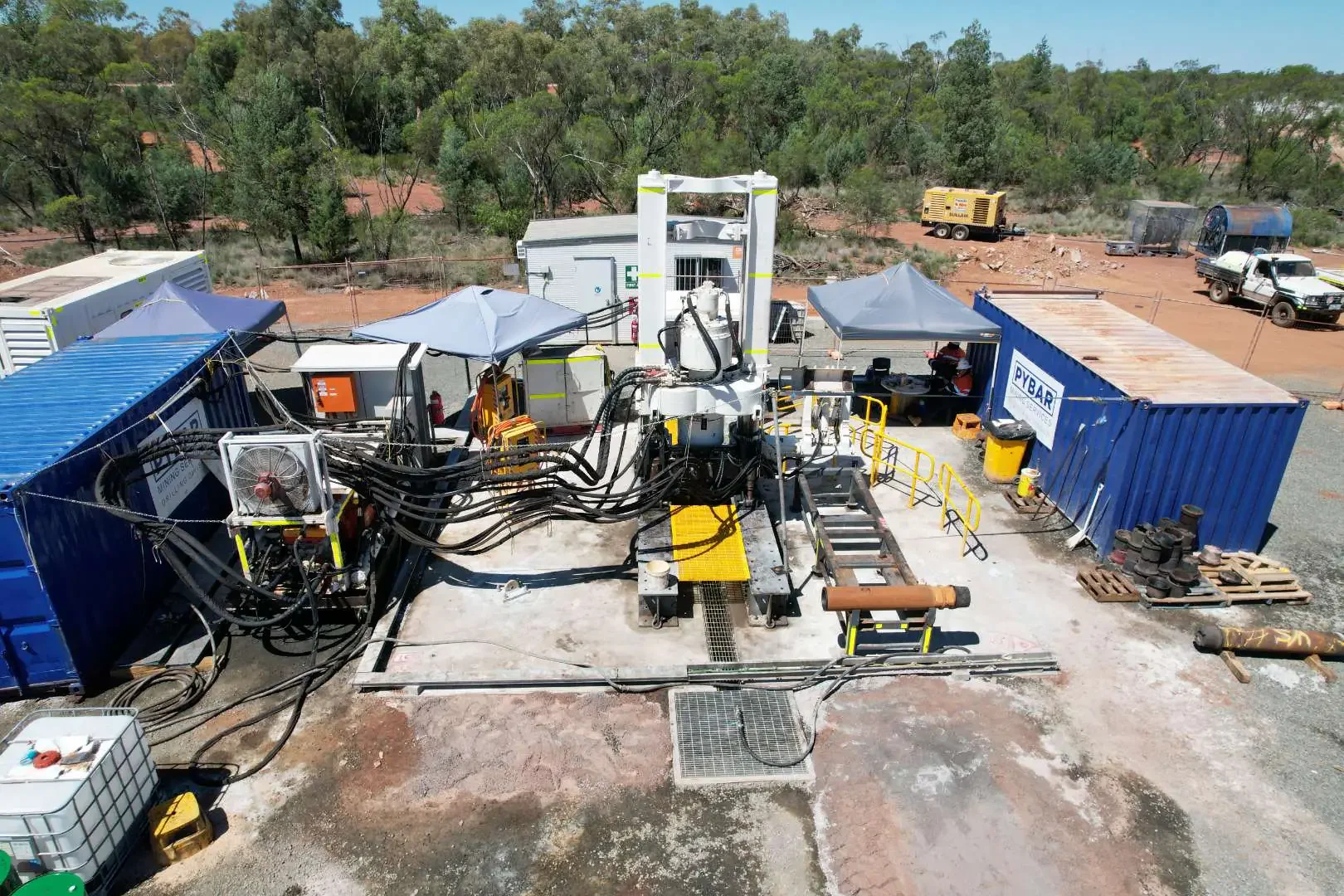 A drilling site setup with heavy machinery and containers in a desert landscape with trees in the background.