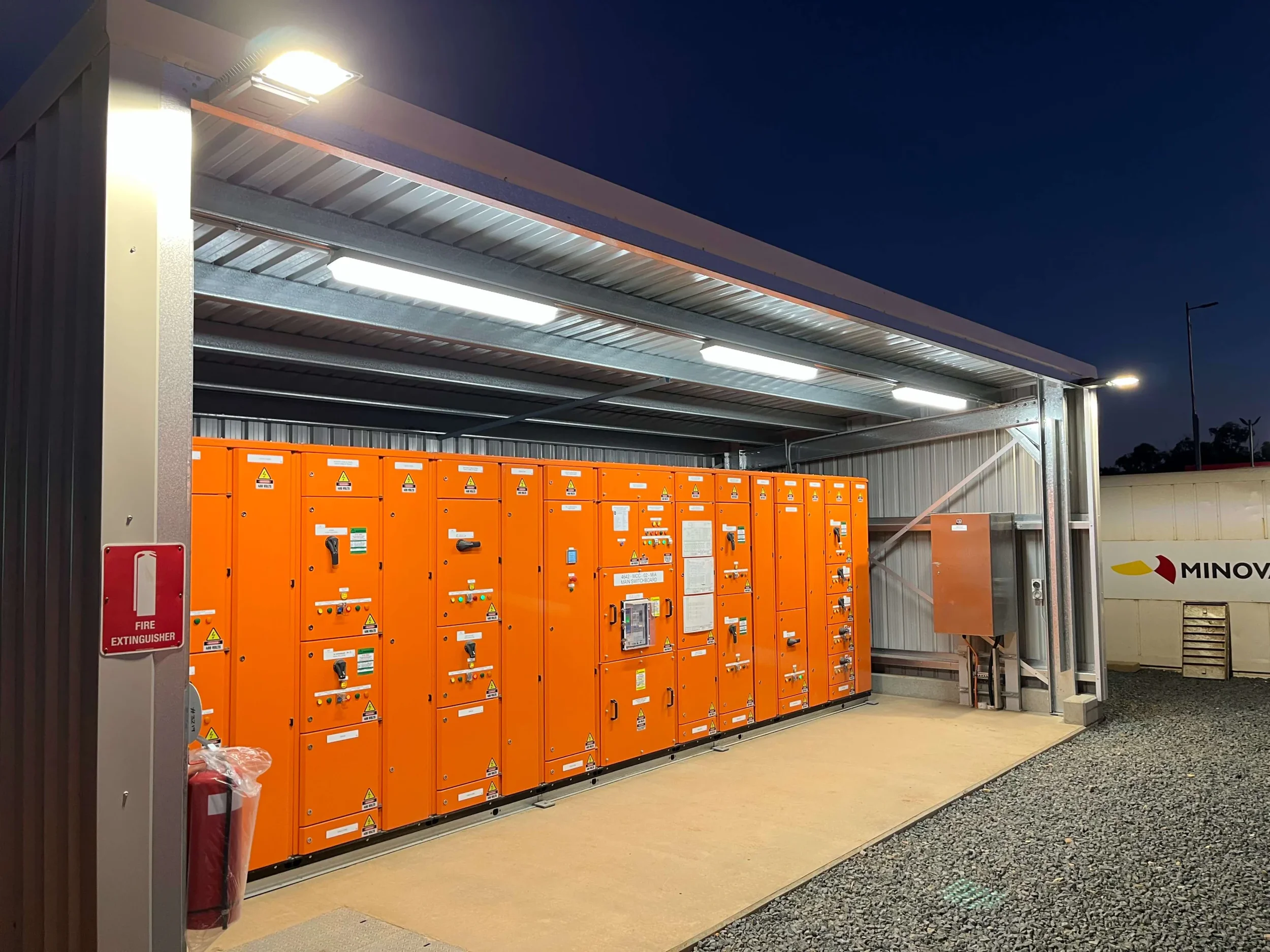 An outdoor electrical substation with orange cabinets, metal framework, and bright LED lighting, set against a clear evening sky.