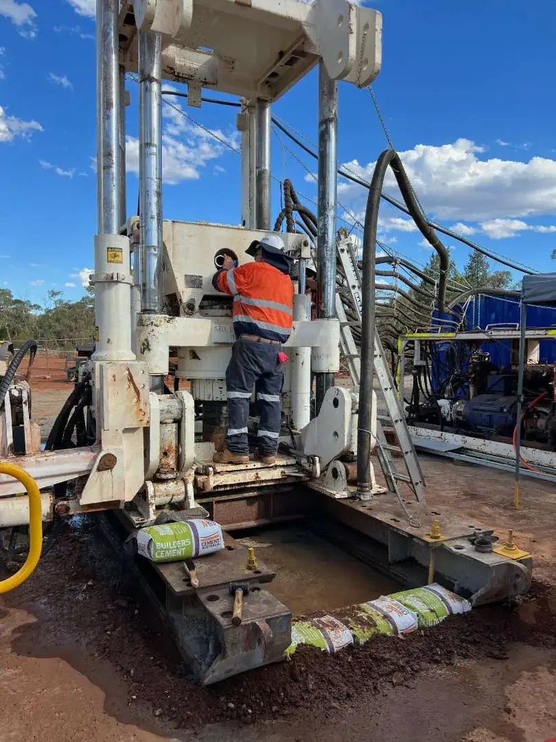 A worker operating a large, white earth-moving machine at a construction site on a sunny day with a clear blue sky and some clouds. The machine is on dirt, with bags of cement nearby, and the worker is wearing safety gear including a helmet and a high-visibility orange vest.