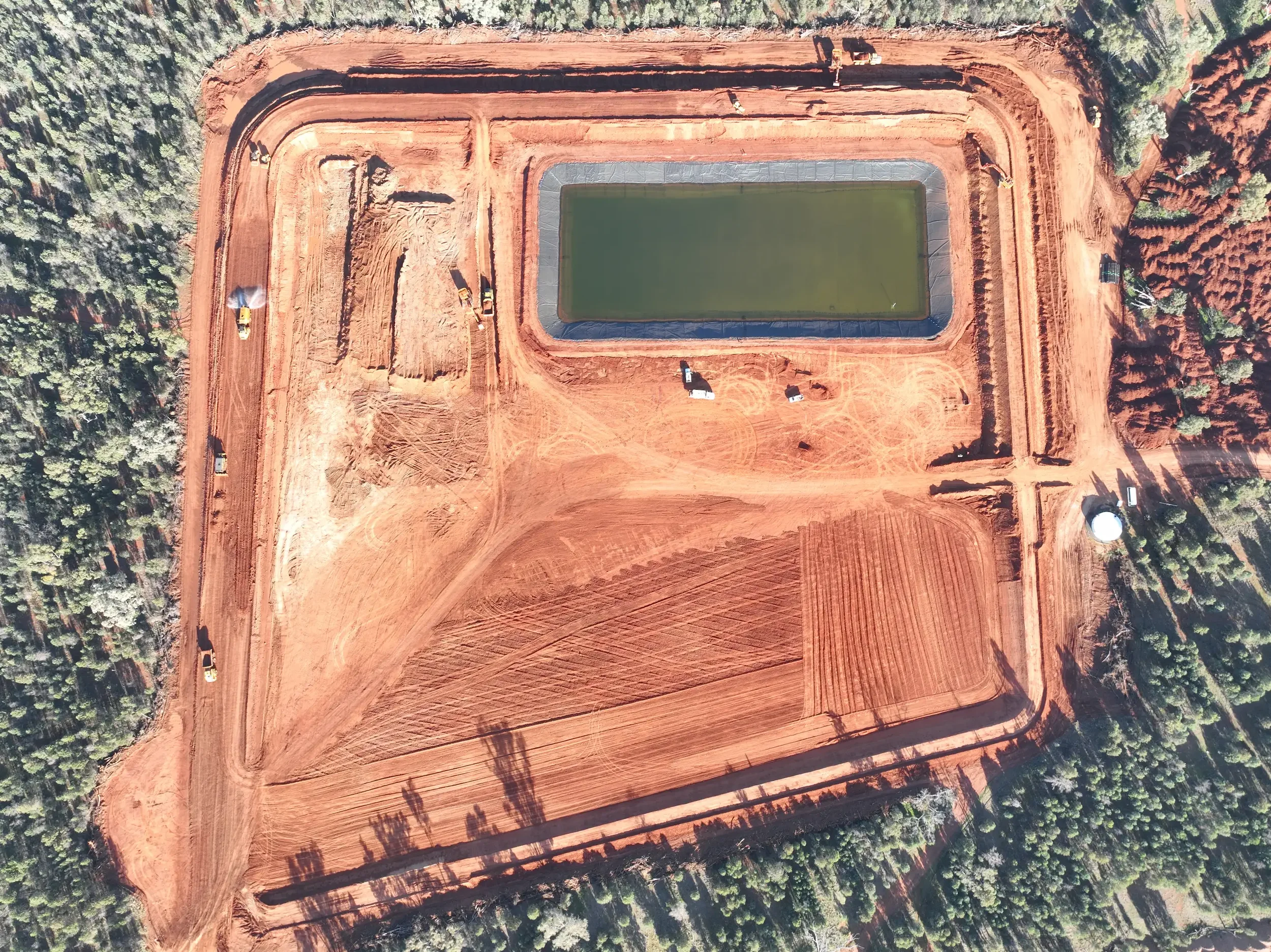 Aerial view of a construction site with a rectangular pond, surrounded by red soil, with construction vehicles and machinery working on the site, and a forested area around the perimeter.