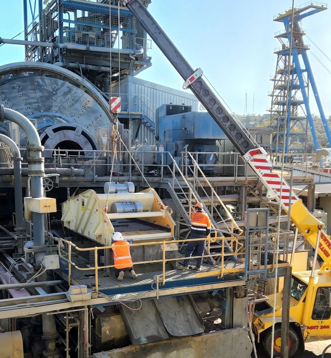 Workers in orange safety vests and white helmets operate machinery on a large industrial site with metal structures and a crane.