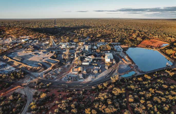 Aerial view of a small town with buildings, roads, a water body, and surrounding forested area at sunset.