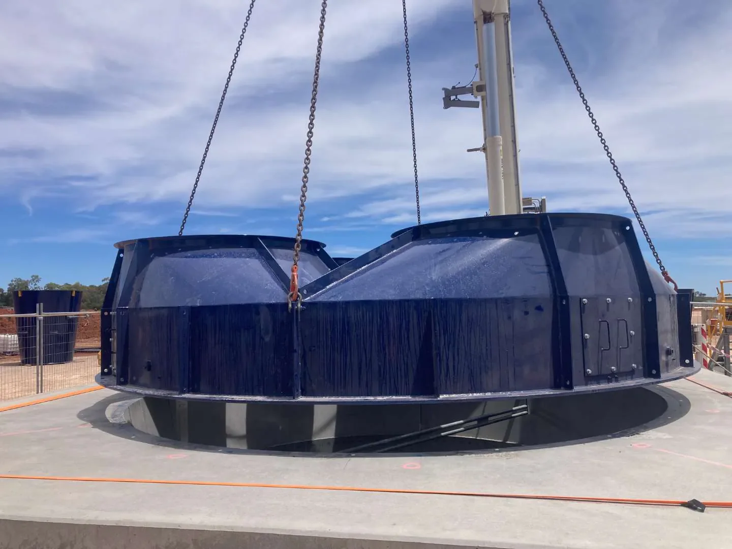 Large blue circular structure with chains hanging from a crane or support, outdoors under a blue sky with some clouds, on a concrete platform.
