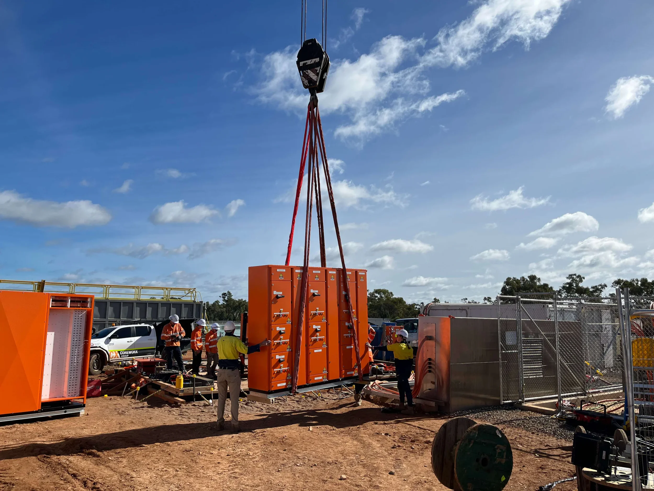 Workers on an electrical construction site assembling large outdoor electrical cabinets using a crane.