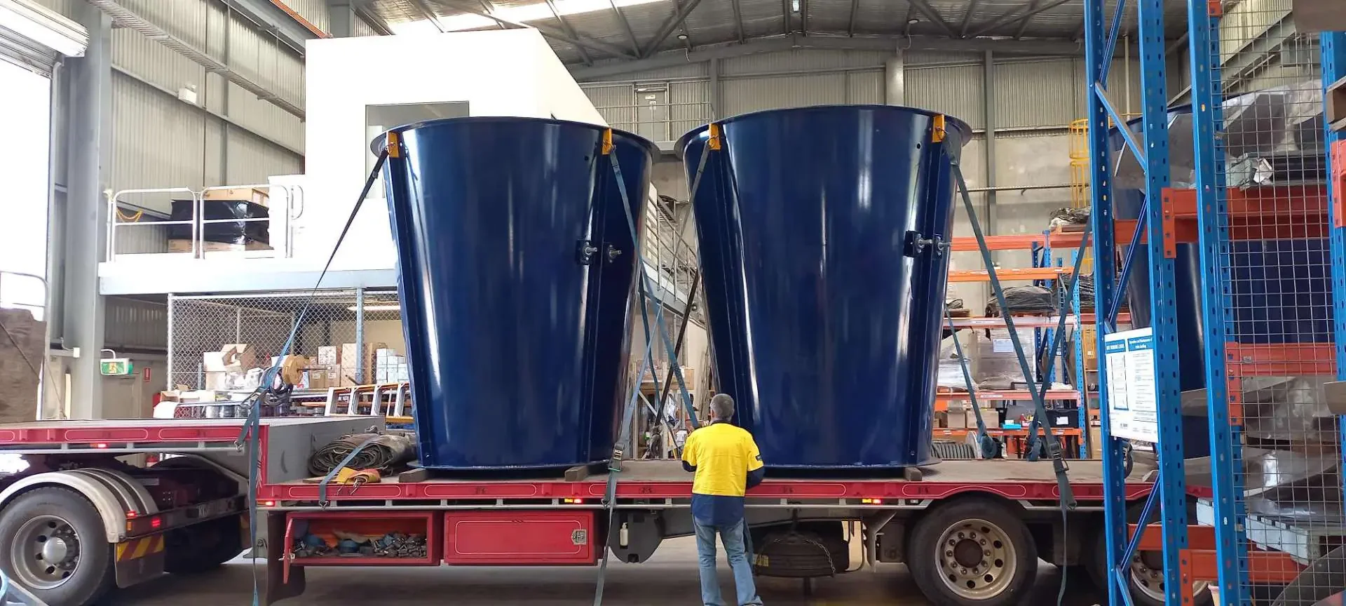Two large blue industrial bins are secured to a flatbed trailer inside a warehouse. A person in a yellow shirt is standing nearby, inspecting the bins.