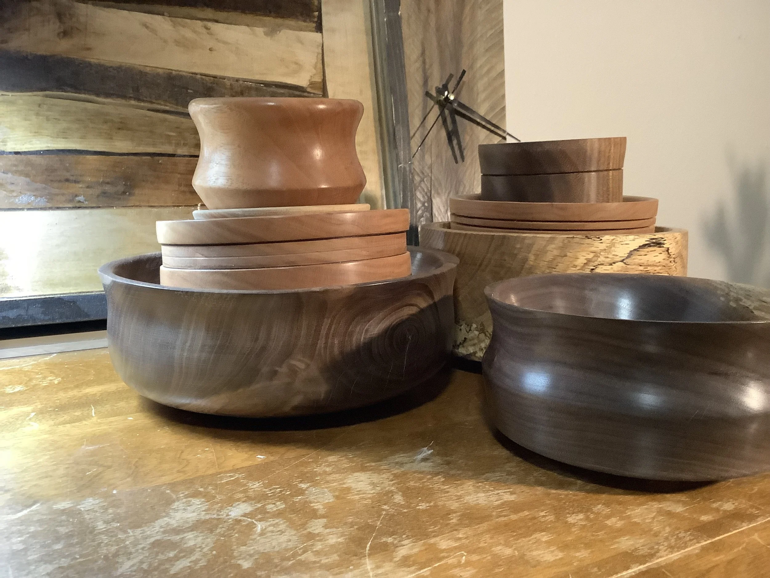 Several wooden bowls and plates stacked on a wooden surface, with a clock and a decorative wall in the background.