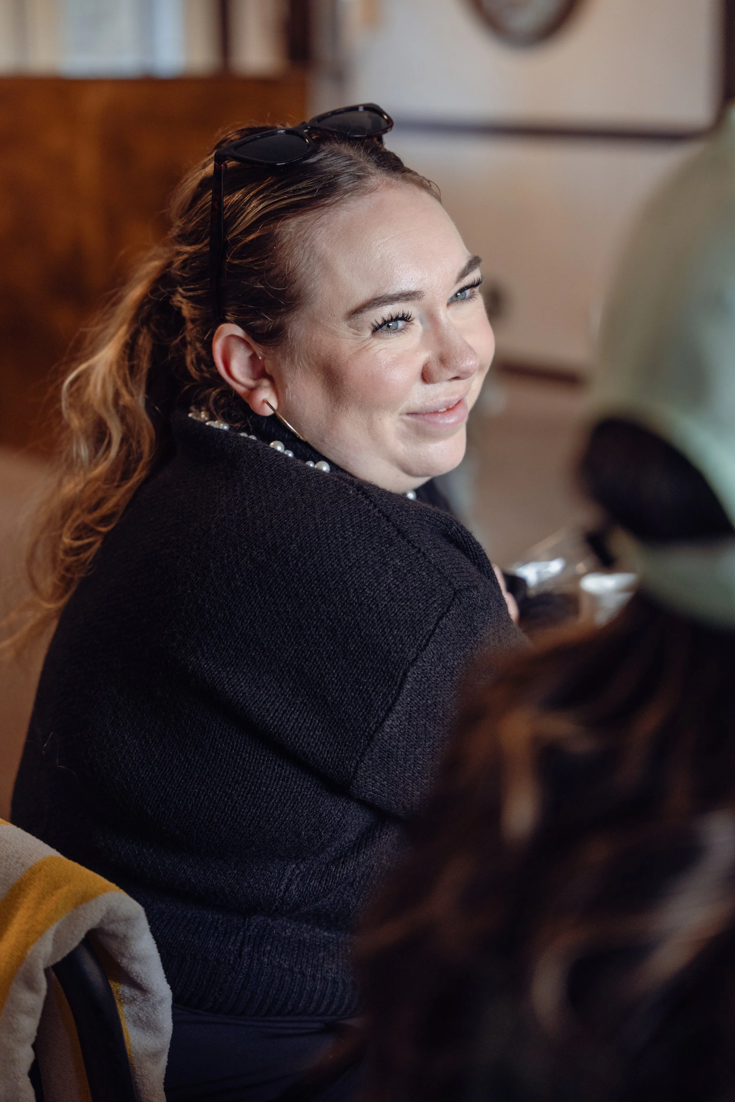 A woman with long, wavy brown hair, sunglasses on her head, wearing a black sweater and a pearl necklace, smiling and looking to her right in an indoor setting.
