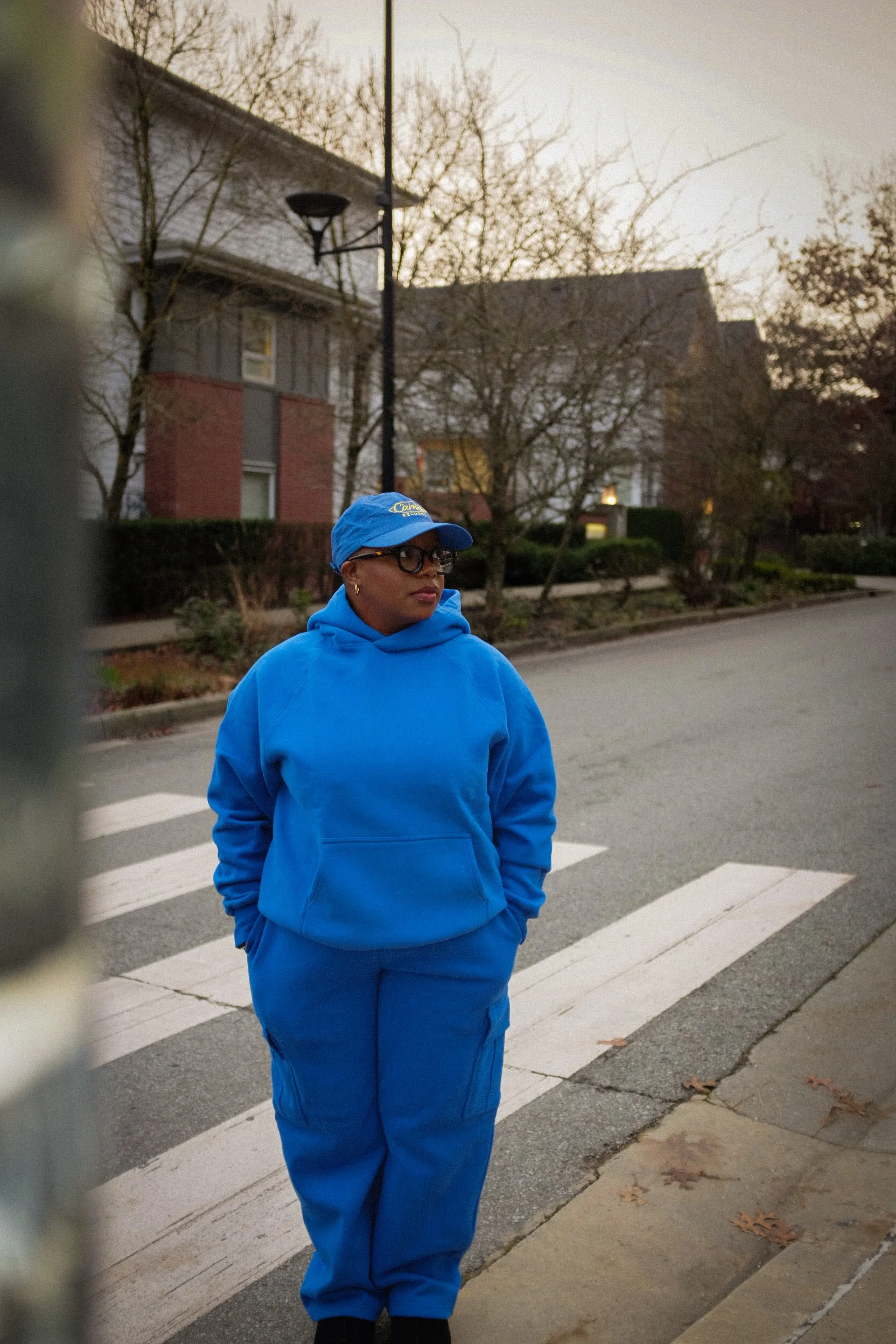 A woman in a blue hoodie, cap, and glasses stands on a crosswalk in a residential neighborhood during dusk.
