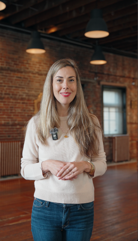 A young woman with long blonde hair wearing a cream sweater and blue jeans, standing in a brick-walled room with large windows and ceiling lights.