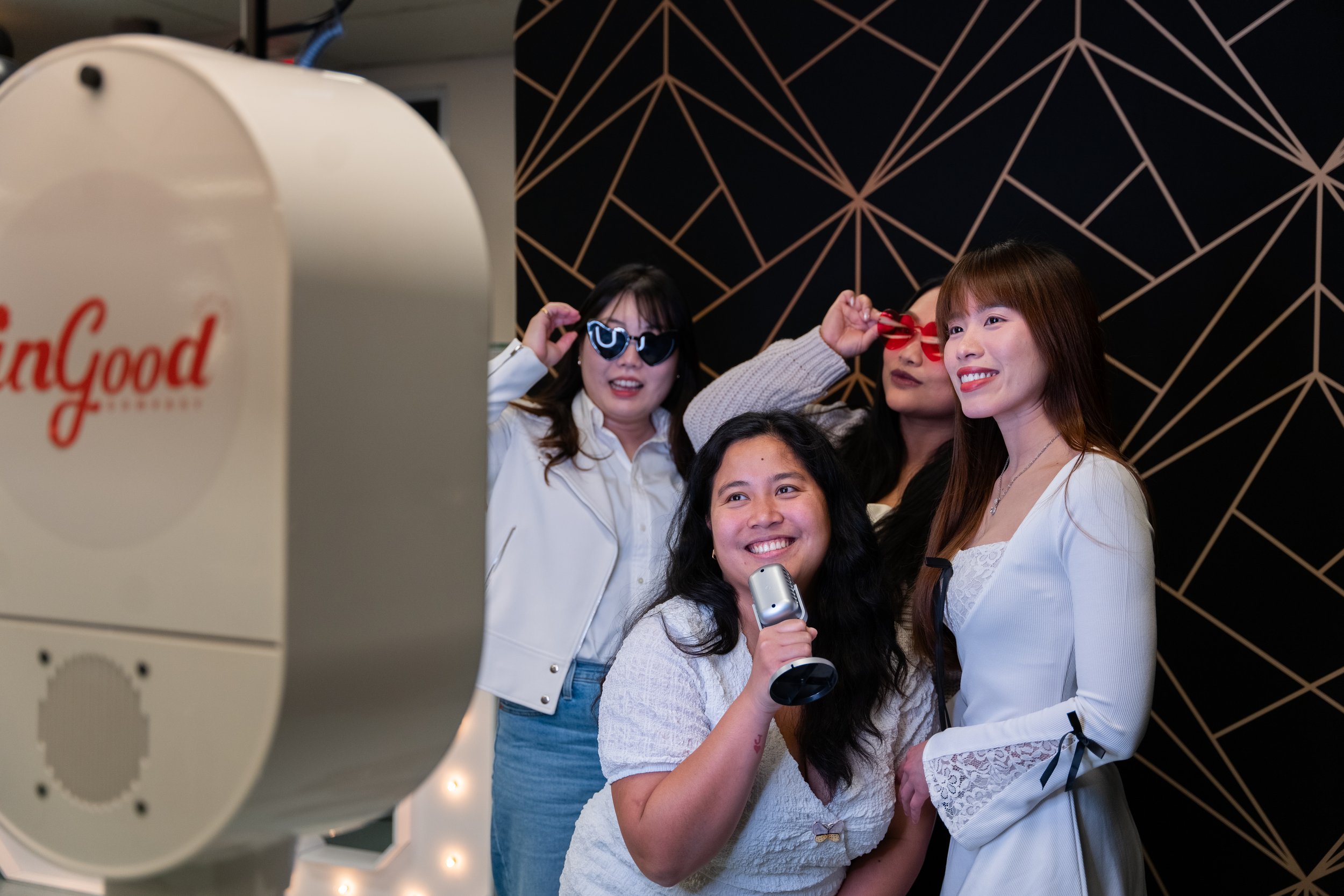 Four women posing for a photo in front of a geometric black and gold patterned backdrop, with a photo booth on the left side of the image.