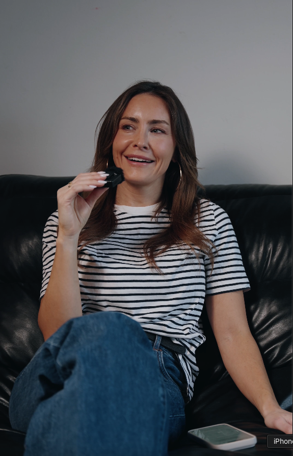 A woman with brown hair, wearing a white and black striped t-shirt, sitting on a black leather couch, holding a TV remote, and smiling while watching television.