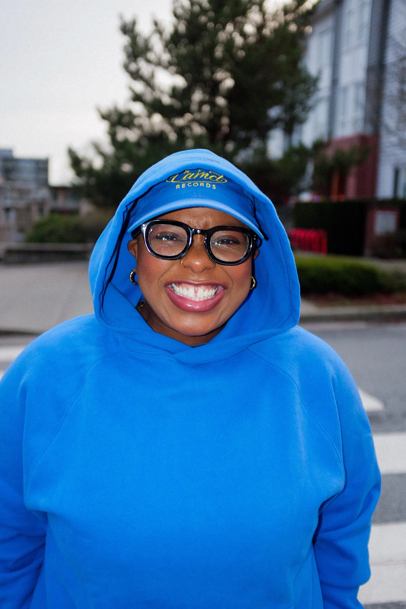 A smiling woman wearing glasses, hoop earrings, and a blue hoodie with the hood over her head, outdoors in an urban setting.