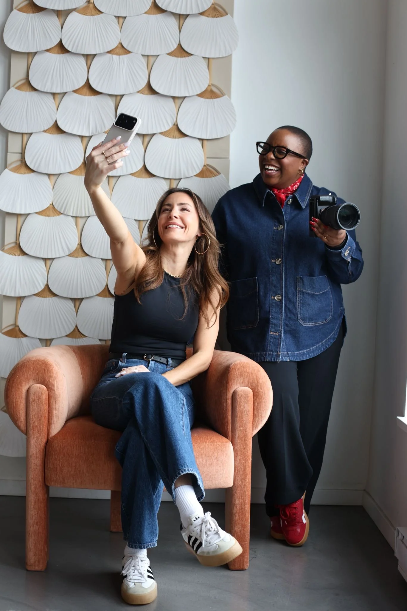 Two women taking a selfie together indoors, one sitting on a pink armchair and the other standing next to her, both smiling and enjoying the moment.