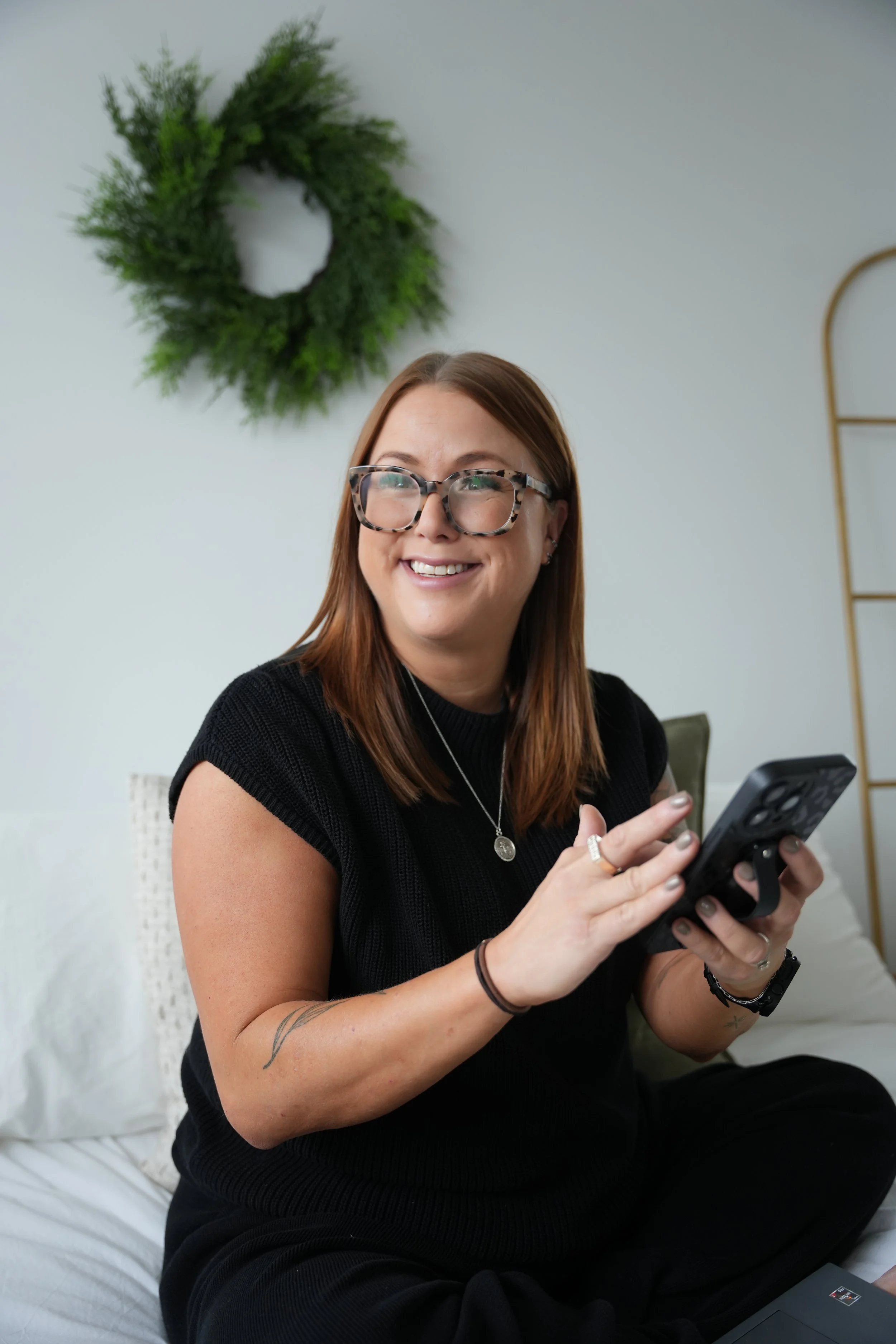 A woman with glasses smiling and holding a smartphone, sitting on a bed in a room with a green wreath on the wall behind her.