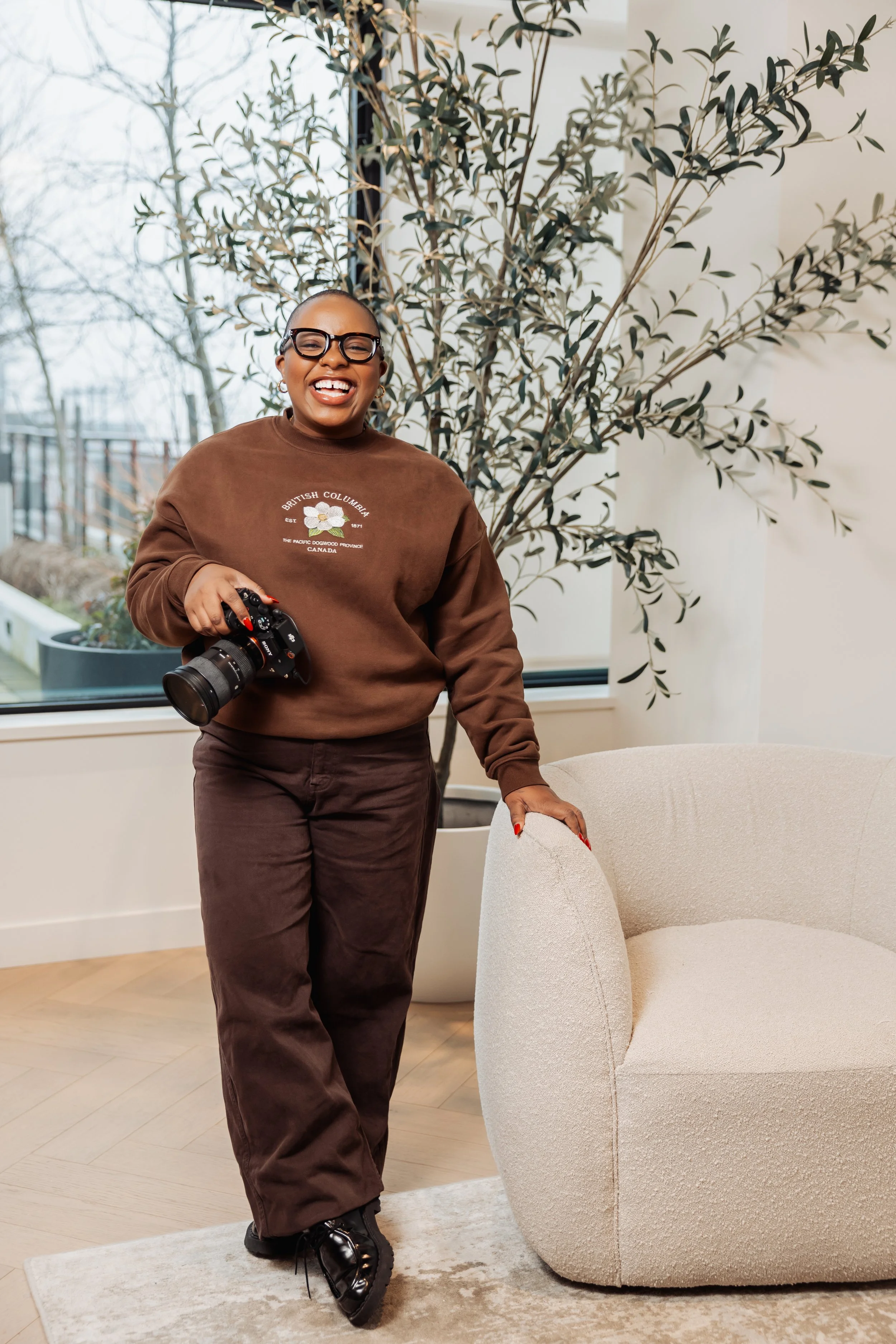 A woman smiling and holding a camera in a modern indoor setting near a beige armchair and a large potted plant.