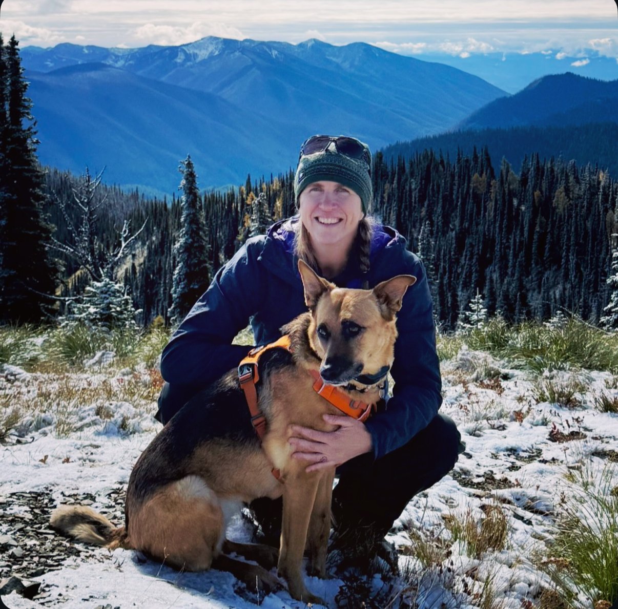 A woman smiling and crouching in a snowy mountainous landscape with a large dog wearing an orange harness.