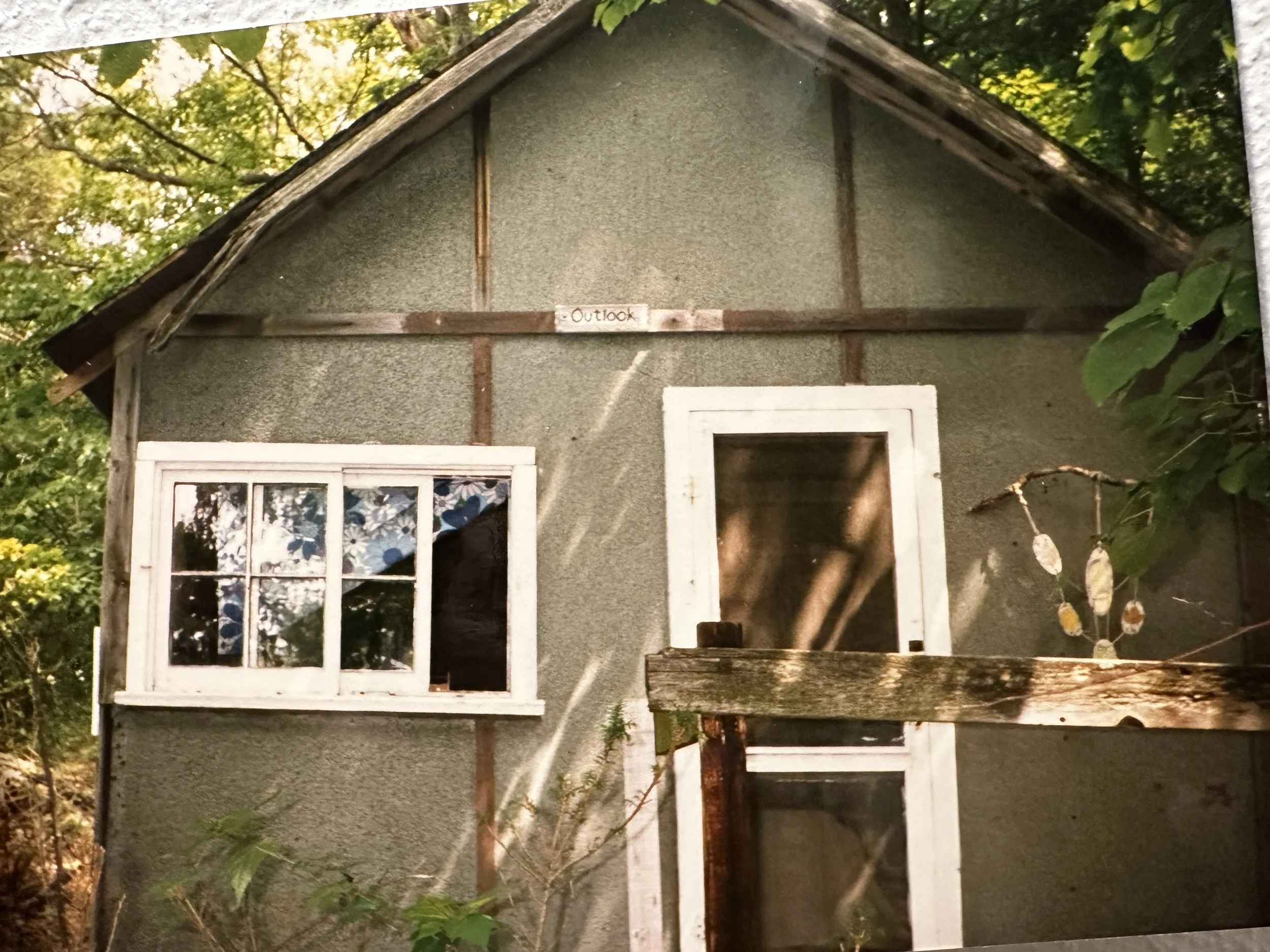 An old, weathered small house or shed with a gray exterior and white-framed windows, surrounded by greenery and trees.