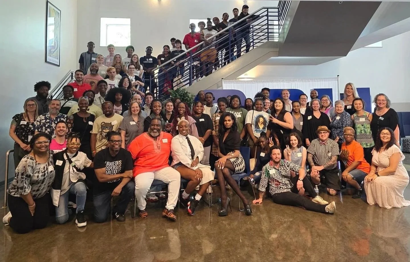 Large diverse group of men and women of various ages gathered for a group photo indoors, with some sitting and others standing on a staircase and in front of a white backdrop.