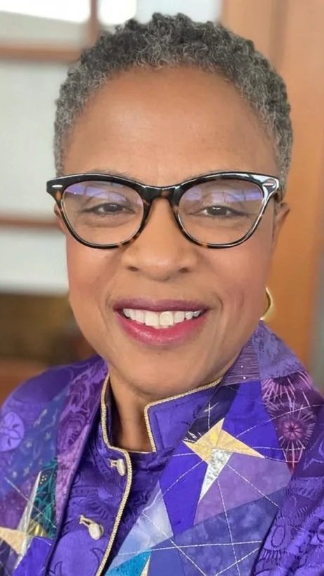 A woman with short gray hair, wearing glasses, a purple jacket with a star pattern, and smiling.