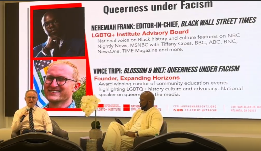 Two men sitting in chairs engaged in a discussion at a panel event. A large presentation slide behind them displays titles and images of two individuals, Nehemiah Frank and Vince Tripi, with their roles and achievements related to LGBTQ+ history and culture activism.