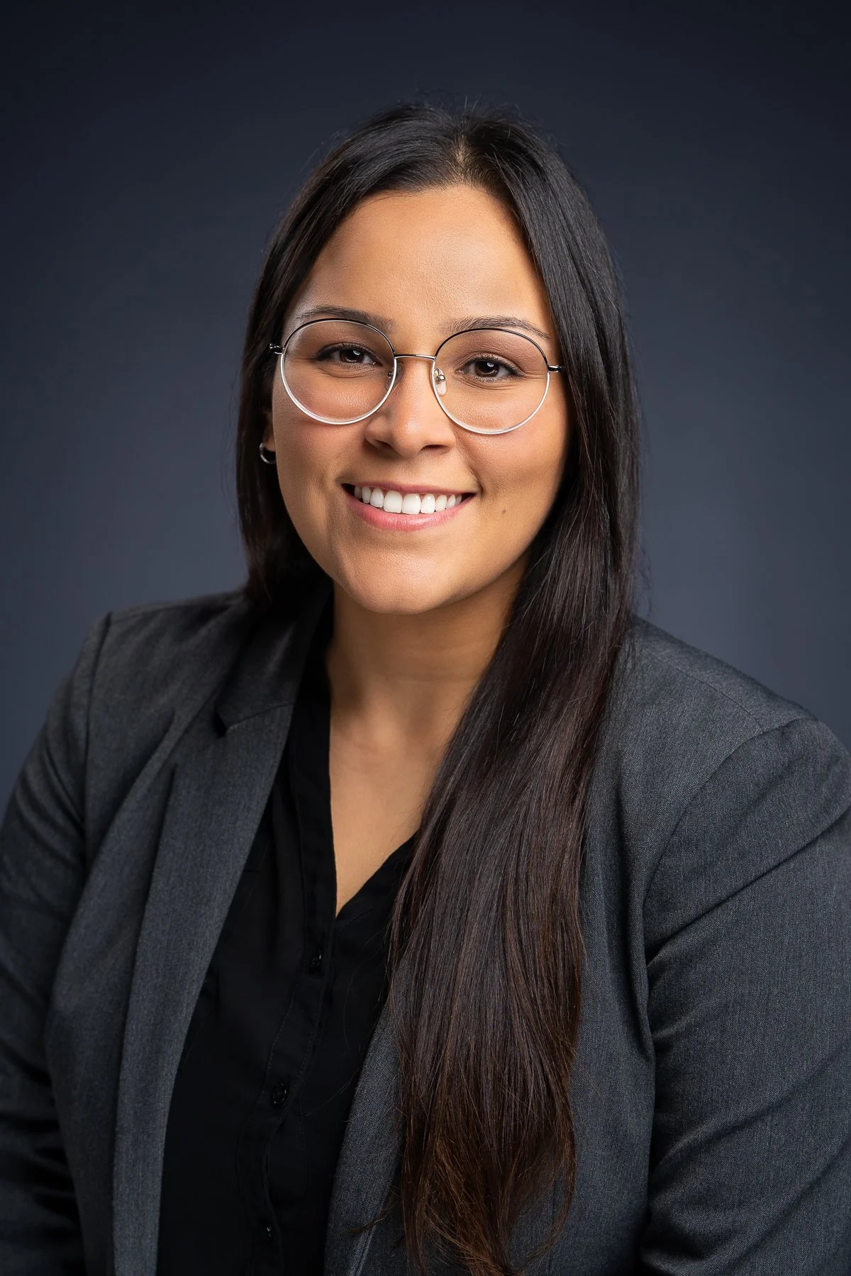 A professional woman with long dark hair, glasses, and a gray blazer smiling at the camera against a dark background.