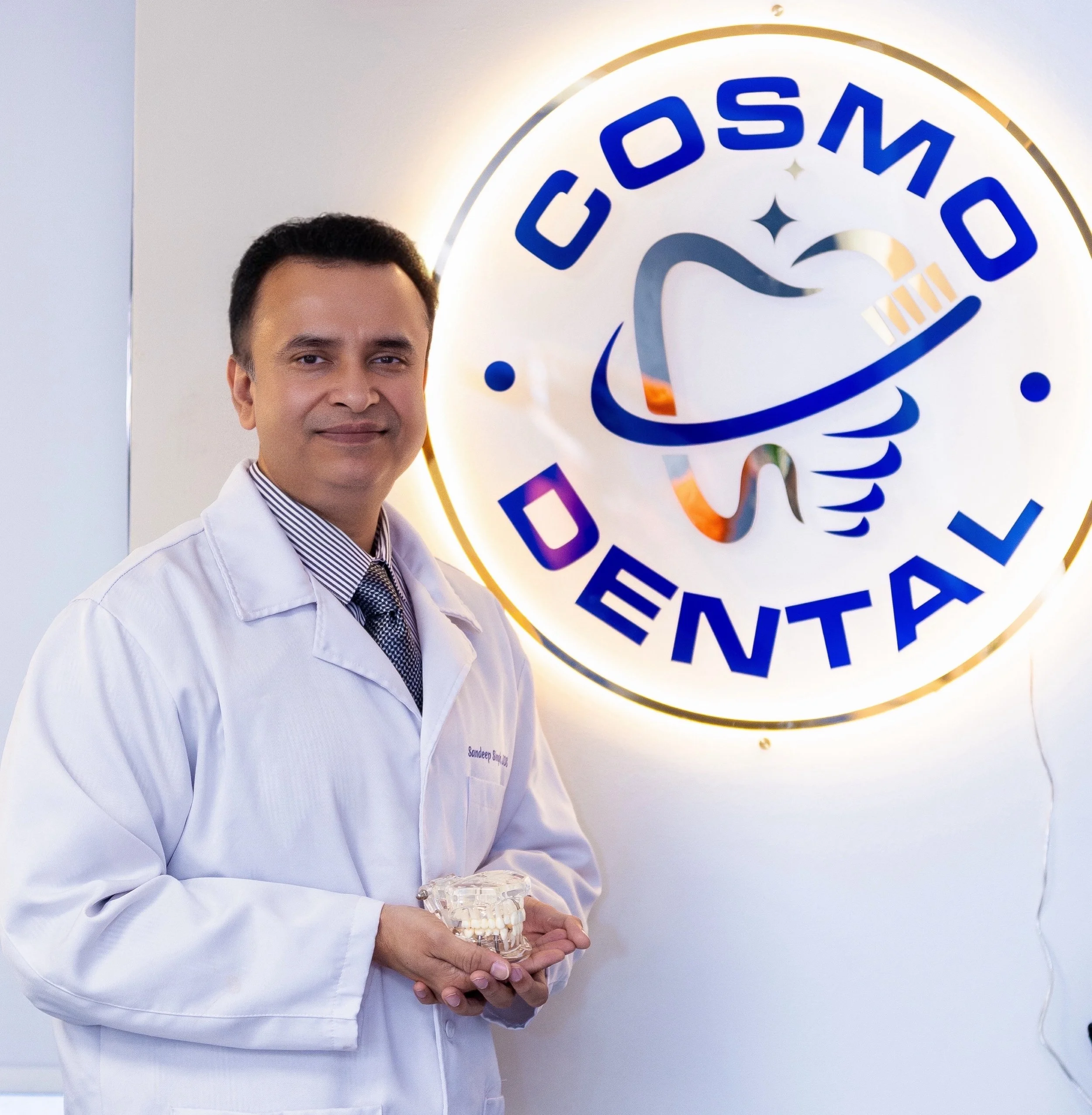 Dr. Singh in a white coat holding a dental model standing in front of a sign that says 'Cosmo Dental' with a tooth and dental tools logo.