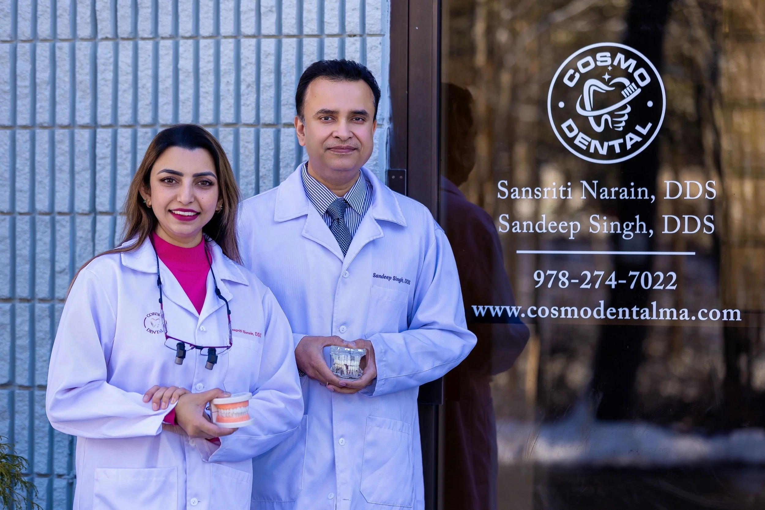 Two dental professionals standing outside a dental clinic, holding dental models. The woman on the left wears a white coat and pink shirt, smiling. The man on the right wears a white coat and tie, holding a dental model. The clinic sign on the window displays the name Sansriti Narain, DDS, Sandeep Singh, DDS, contact number, and website.
