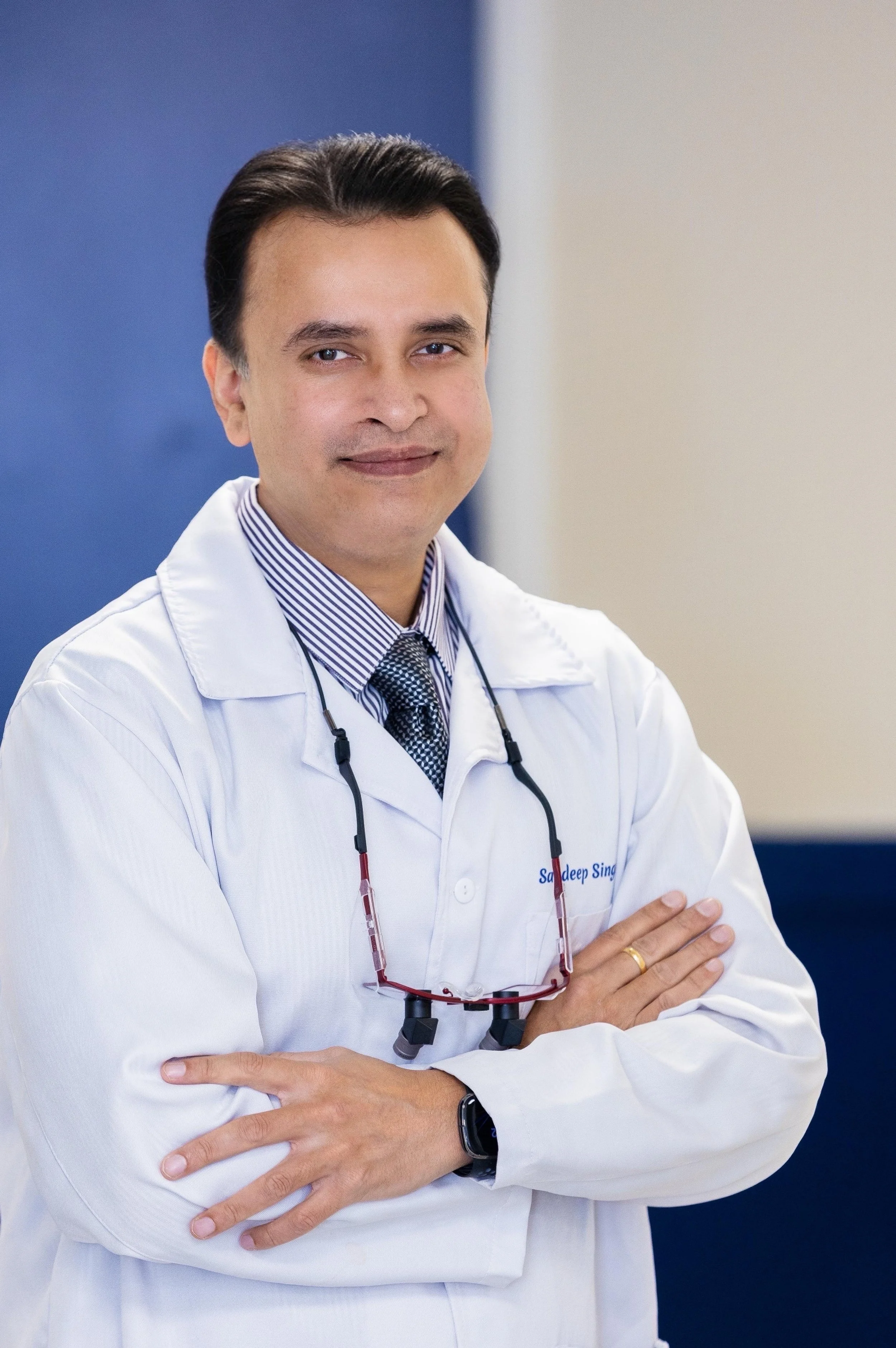 Male dentist, Dr. Singh, with dark hair, wearing a white lab coat, striped shirt, and tie, standing confidently with arms crossed in a medical office.
