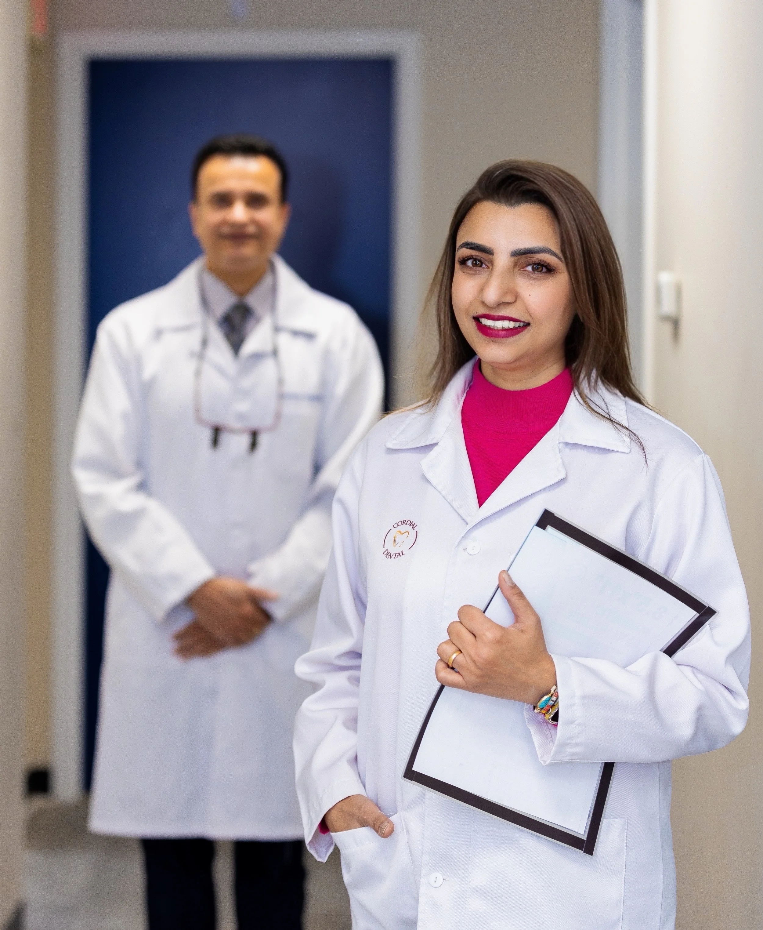 Smiling female dentist, Dr. Narain, holding a clipboard, standing in front of male colleague, Dr. Singh, both dressed in white coats outside a medical office.
