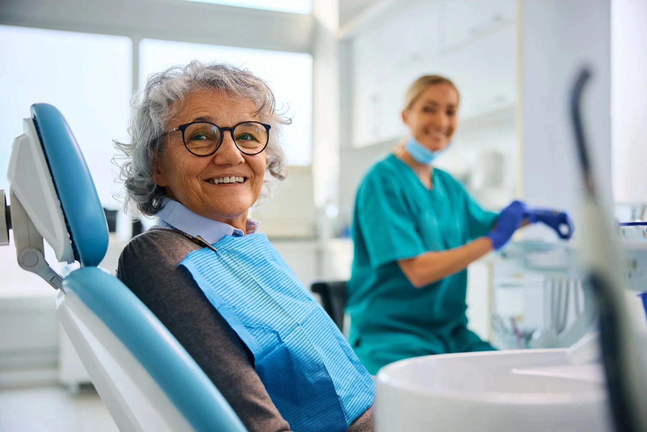 An elderly woman with glasses sitting in a dental chair, smiling, with a dental assistant in scrubs and gloves in the background, in a bright dental clinic.