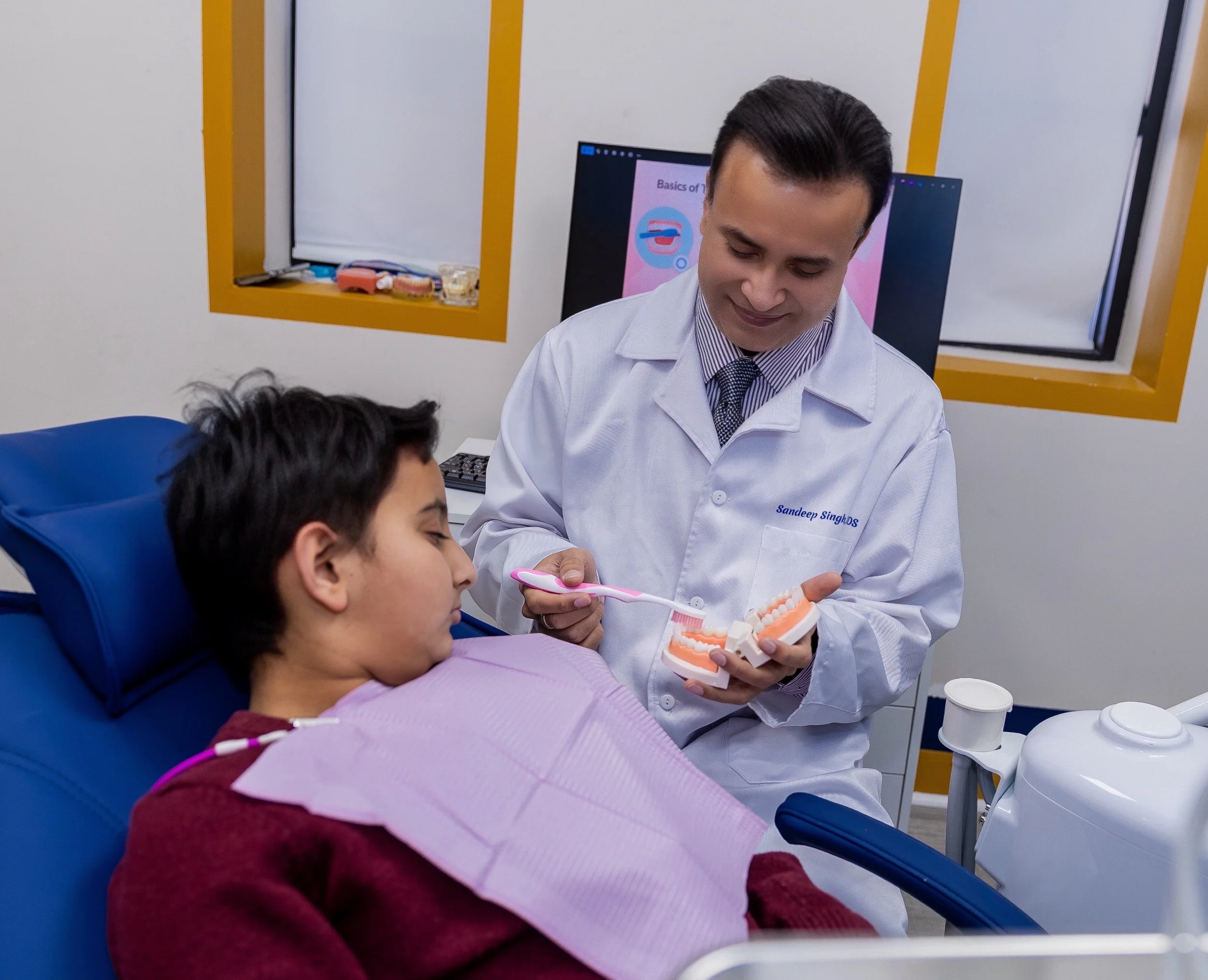 Dr. Singh showing a dental model and toothbrush to a child patient sitting in the dental chair.