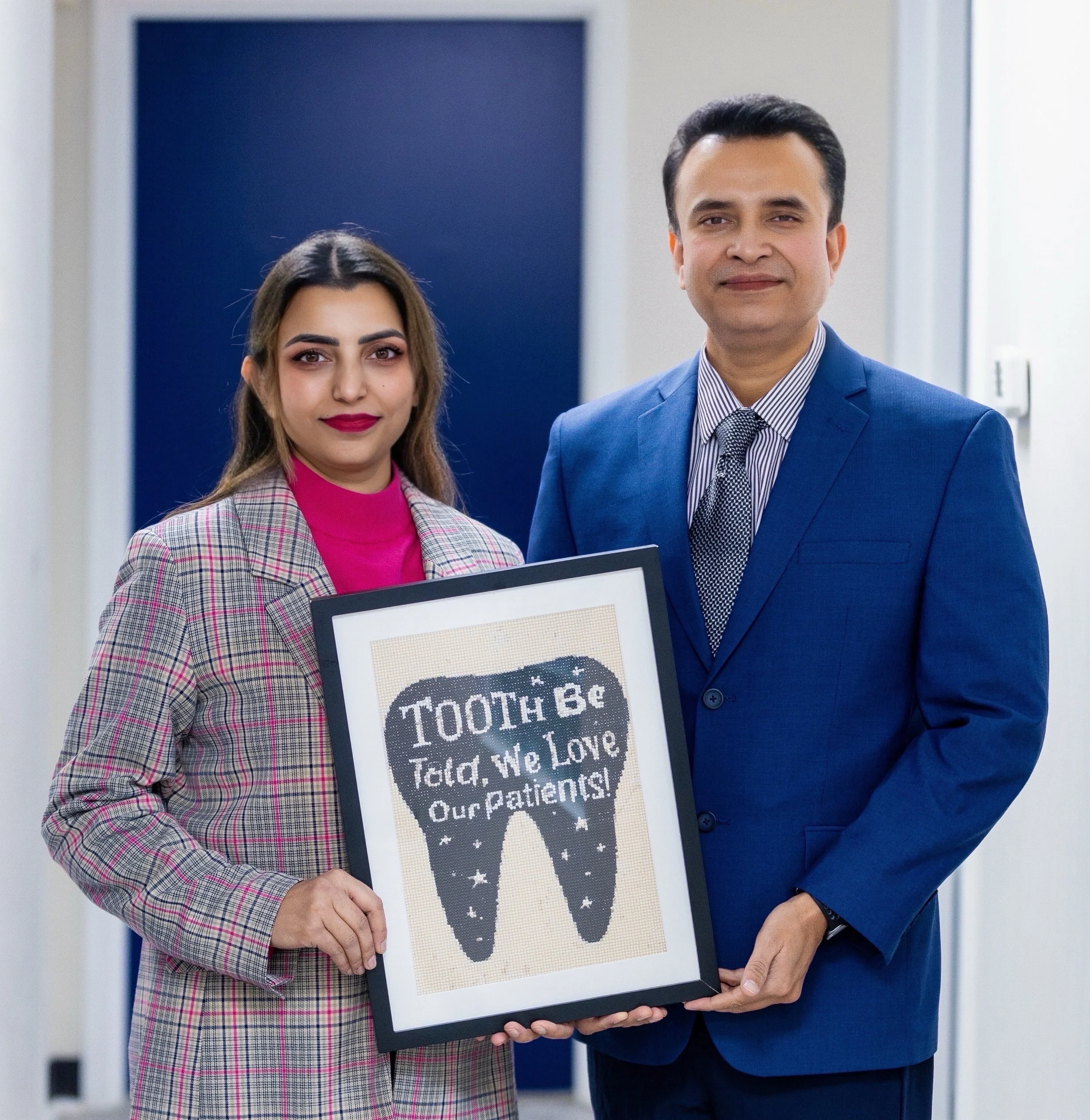 Dr. Narain and Dr. Singh holding a framed cross-stitch art of a tooth with the message 'Tooth be Told, We Love Our Patients'.