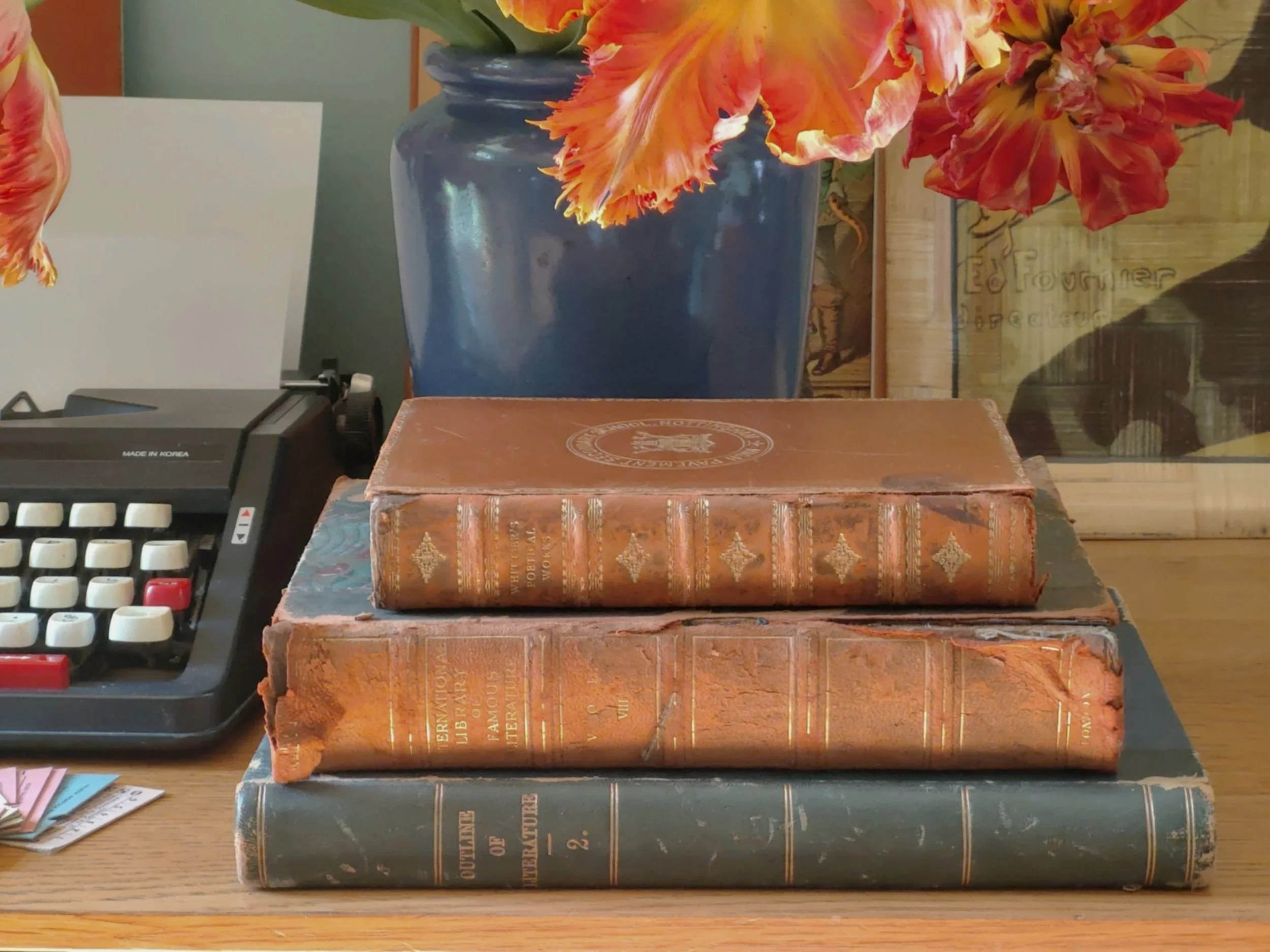 Three stacked antique books on a wooden table with a blue vase of pink and orange flowers and a vintage cash register in the background.