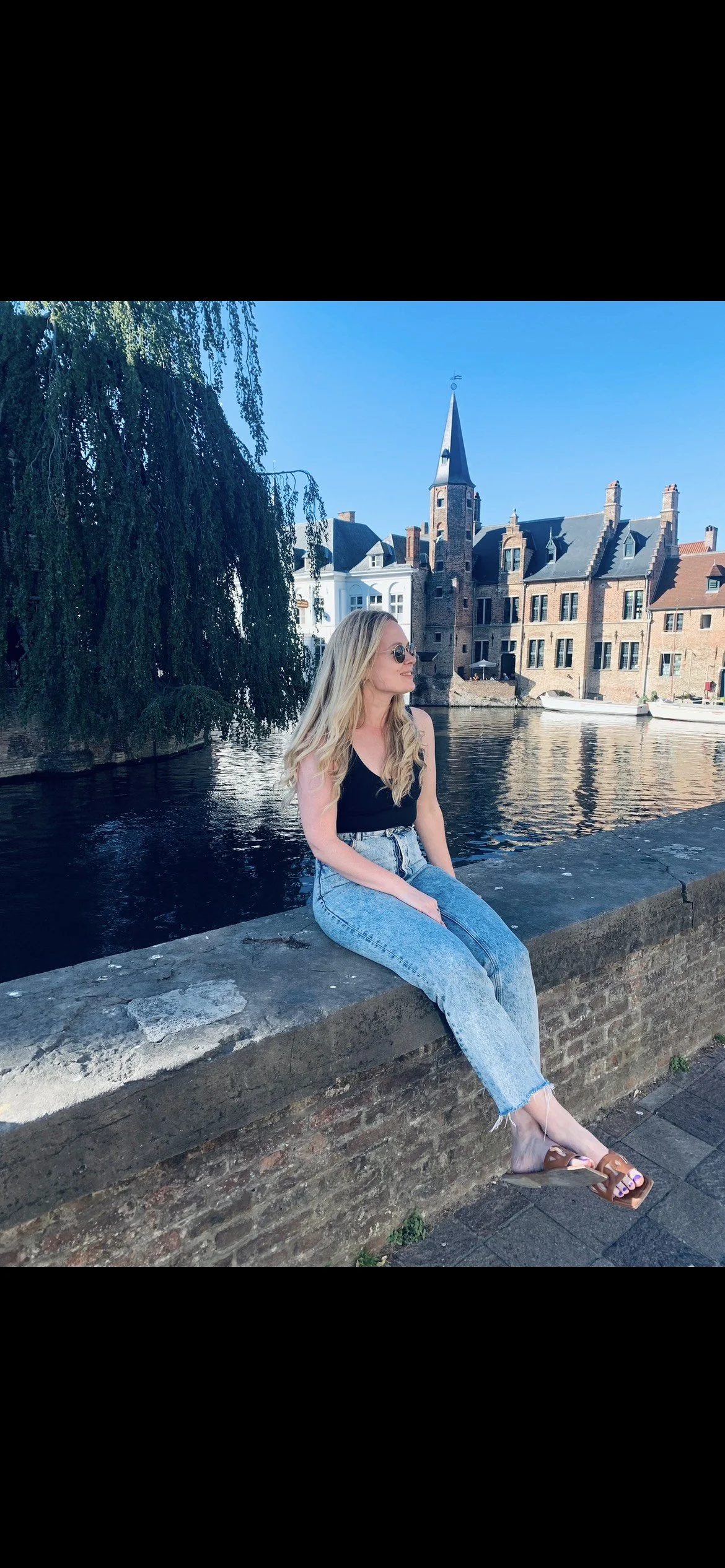 A woman with long blonde hair wearing sunglasses, a black top, and blue jeans sits on a stone ledge by a canal, with historic European-style buildings, a church steeple, and a tree reflecting in the water in the background on a bright, sunny day.