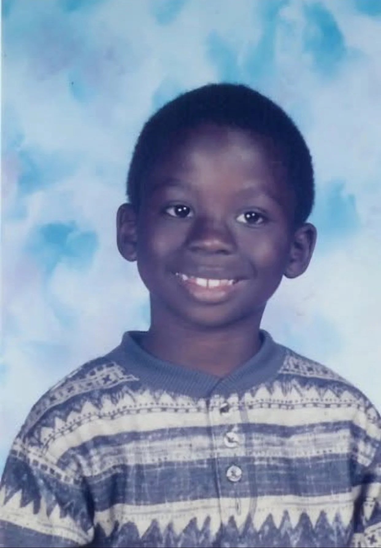 Smiling young boy with short black hair, wearing a patterned sweater, posing for a school portrait against a blue cloudy background.