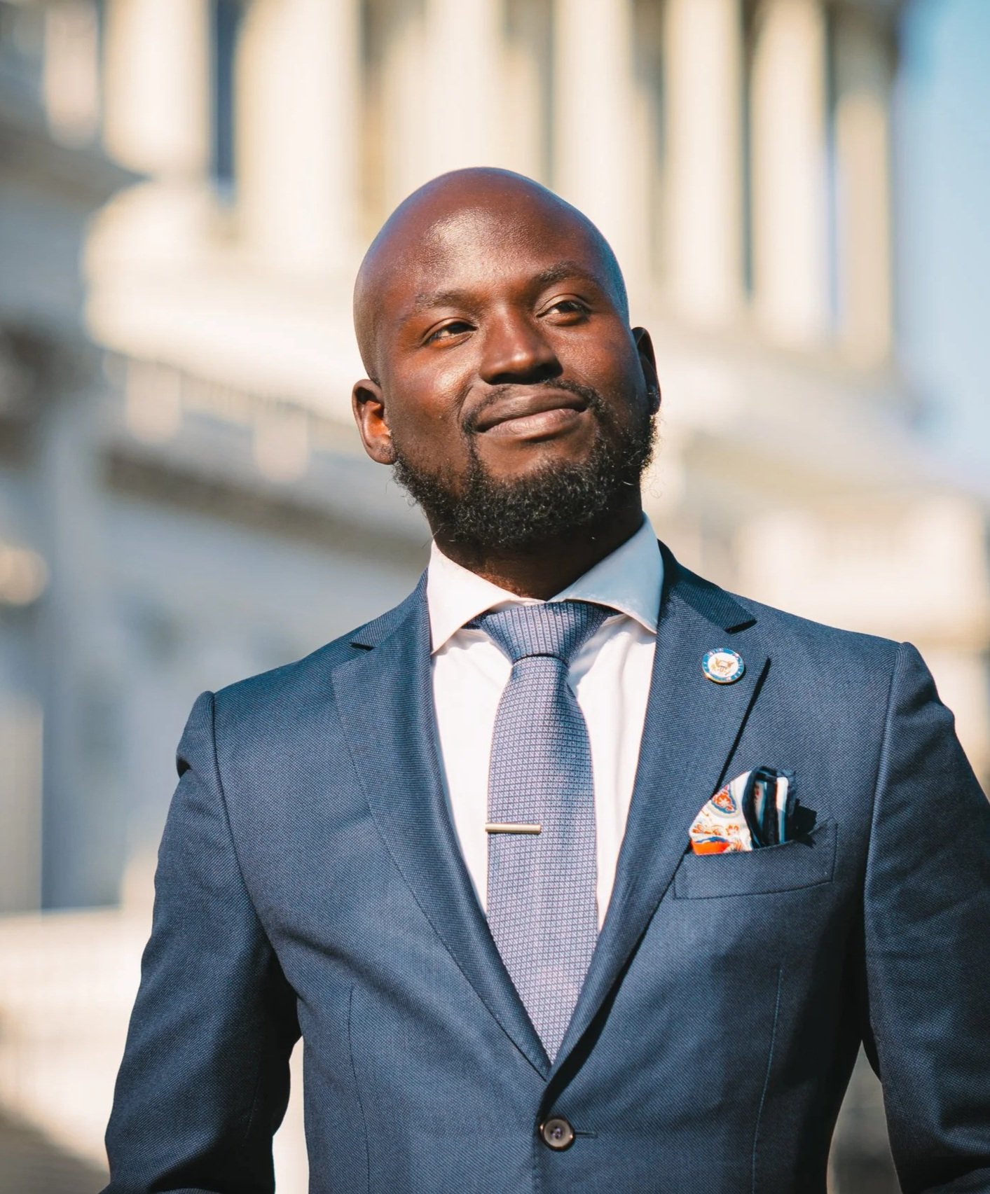 A confident African American man in a navy suit and light blue tie stands outdoors with a blurred cityscape background.
