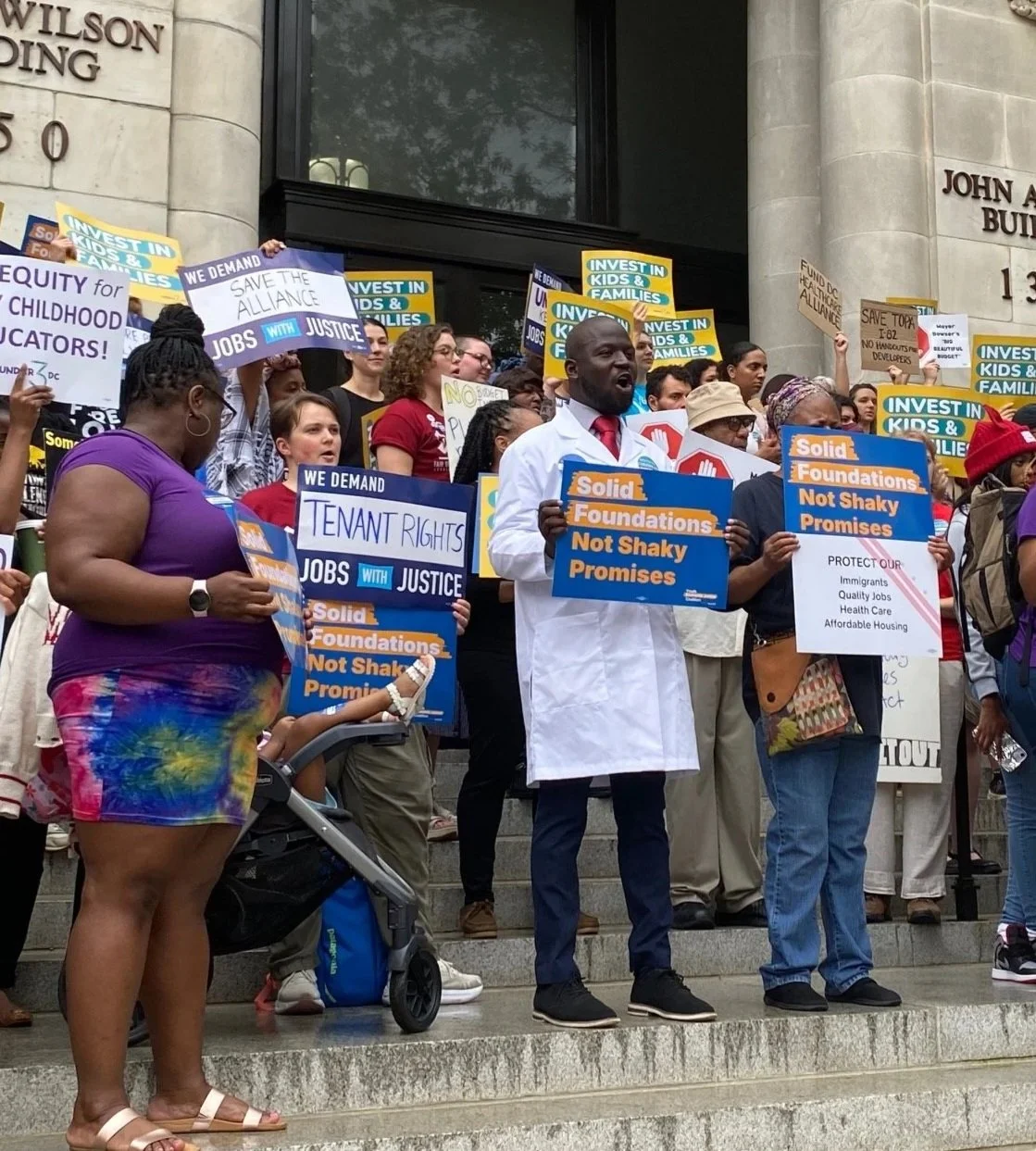 Group of protesters holding signs and banners advocating for tenant rights, jobs, justice, and investment in families outside a government building with stone steps and a sign reading 'Wilson Building.'