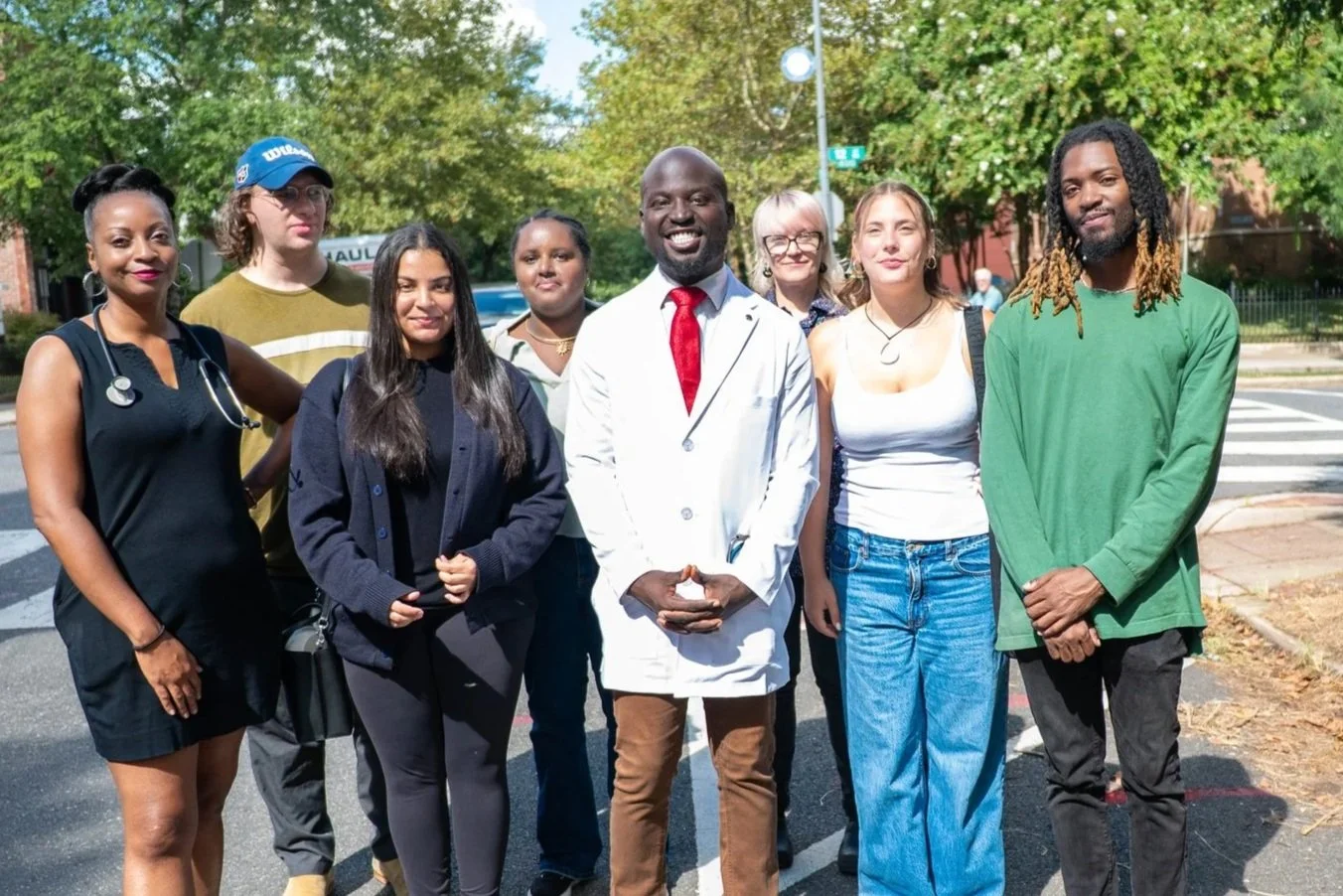 Group of diverse men and women standing outdoors on a sunny day, with a man in a white lab coat and red tie at the center, smiling.
