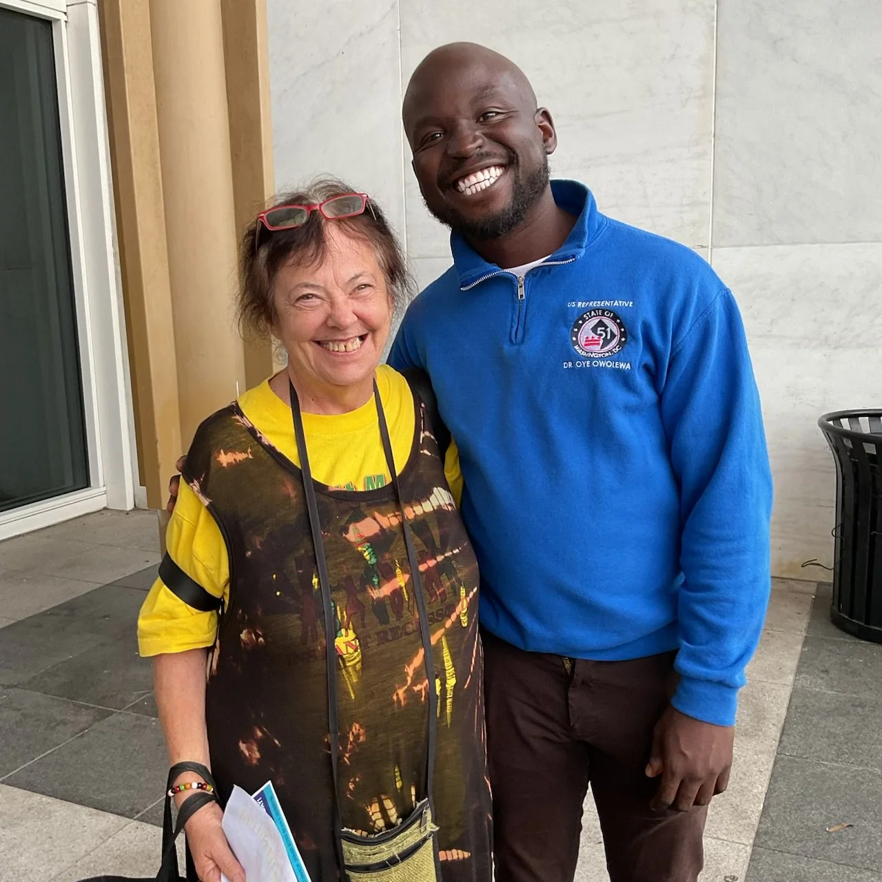 Two smiling people, a woman and a man, standing close together outdoors, with the woman wearing glasses on her head and holding papers, and the man wearing a blue jacket with an emblem for a U.S. representative, in front of a building.