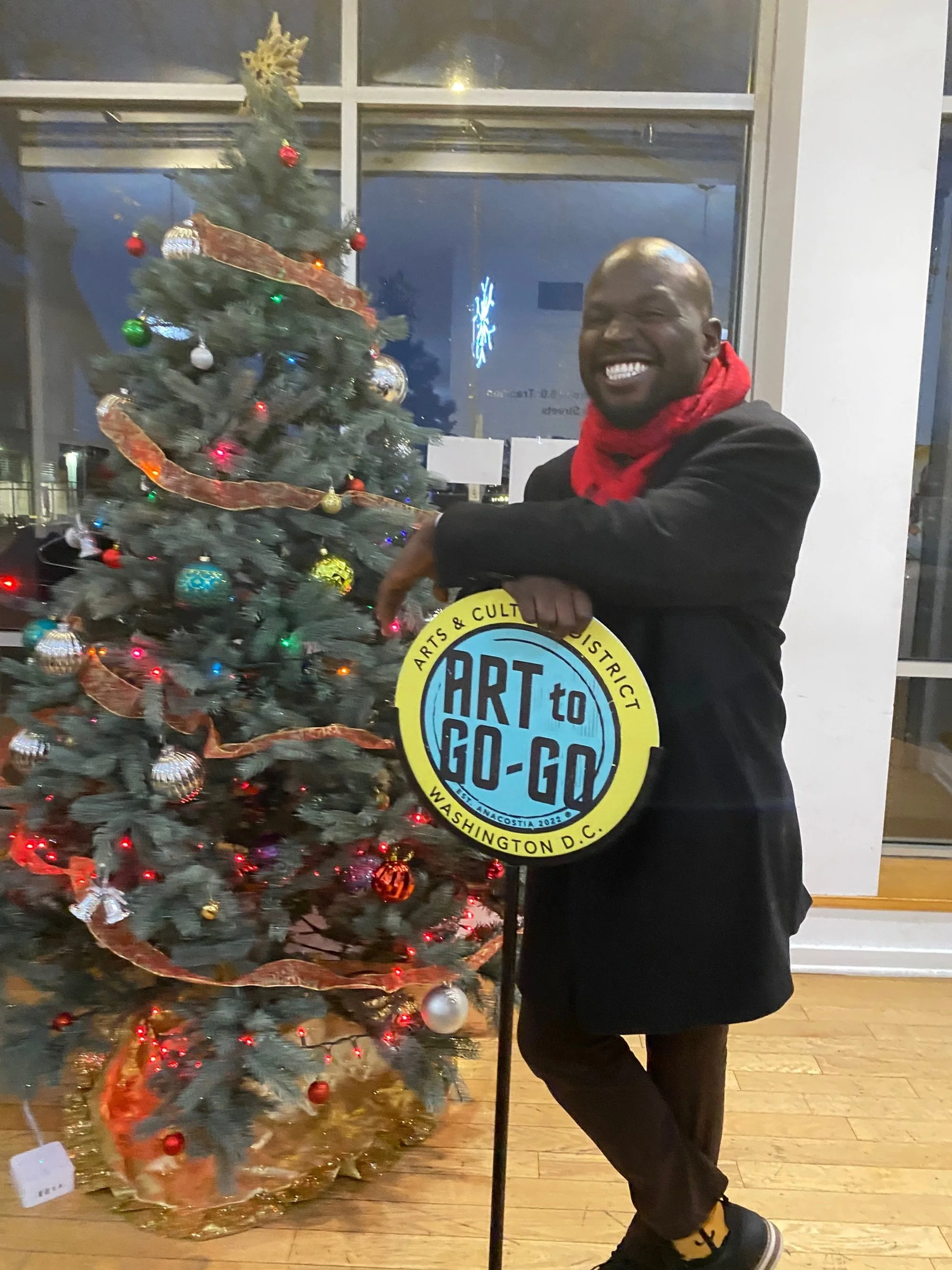 A man standing indoors next to a decorated Christmas tree, holding a sign that reads 'ART TO GO-GO' with a smile on his face.